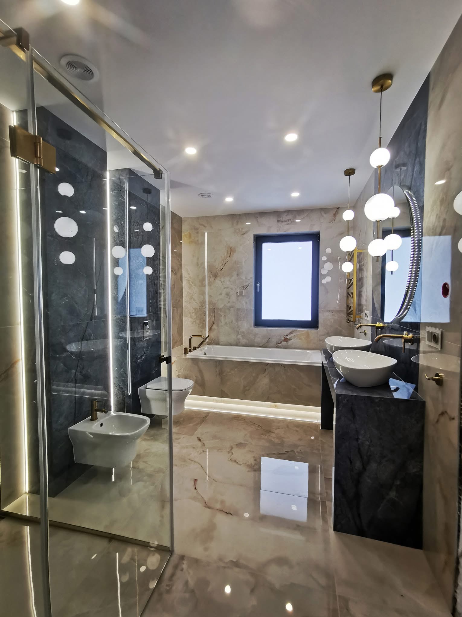 Nowoczesna łazienka z marmurowymi płytkami, szklaną kabiną prysznicową z podświetleniem LED, wiszącą armaturą i okrągłym lustrem podwieszonym na złotych prętach.