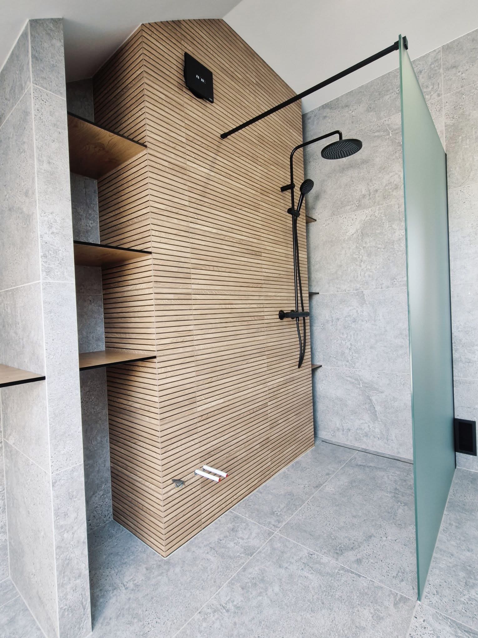 Nowoczesna łazienka z kabiną typu walk-in, drewnianą ścianą z lamelami i półkami. Szara glazura, czarna armatura, minimalistyczny design.