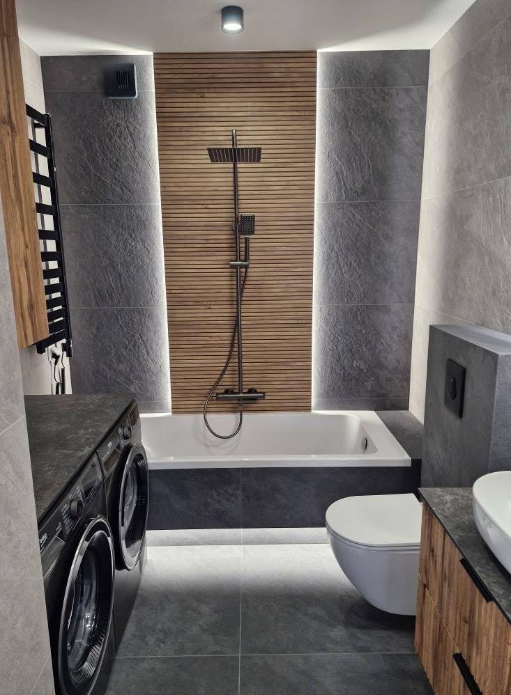 Nowoczesna łazienka z drewnianą ścianą nad wanną, szarą płytką imitującą kamień, podświetleniem LED i czarną armaturą. Widoczna pralka i toaleta.