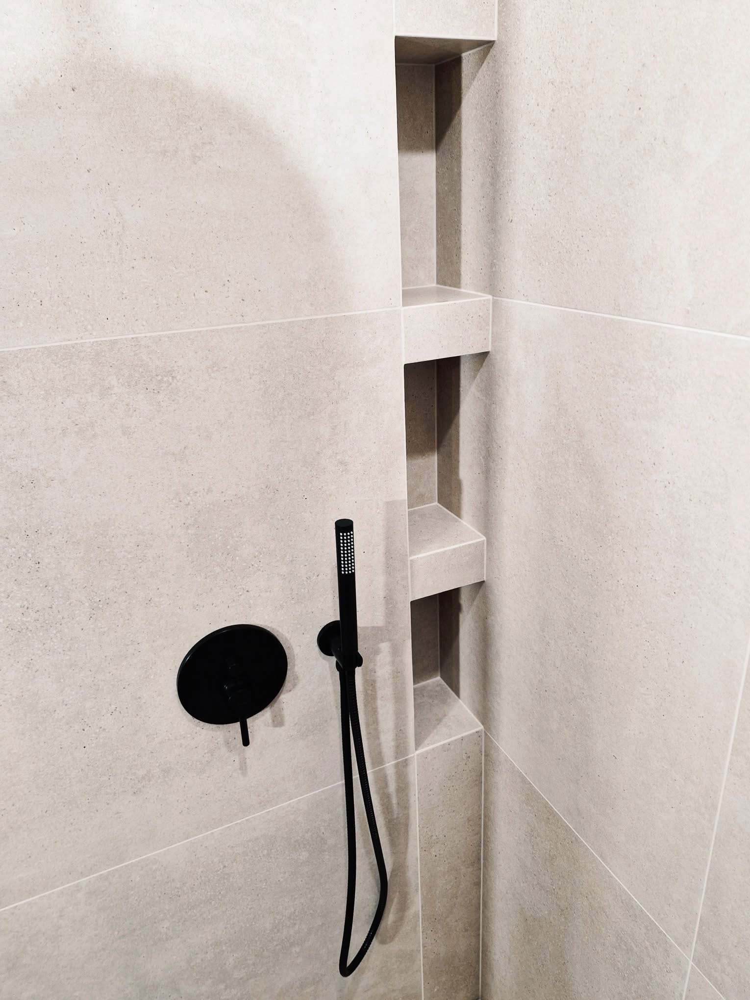 Minimalistyczna kabina prysznicowa z beżowymi płytkami i czarną armaturą, wnęki w ścianie. Nowoczesny design, stonowana kolorystyka, precyzyjne wykończenie.