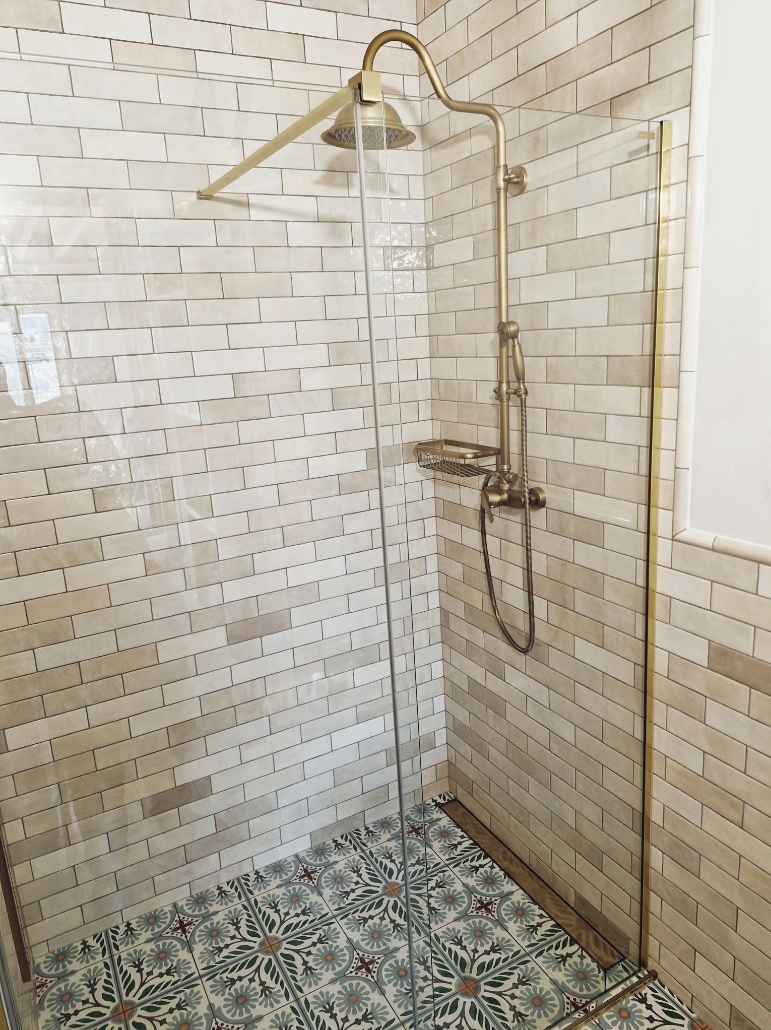 Kabina prysznicowa z beżowymi, cegiełkowymi płytkami i wzorzystą podłogą. Złota armatura i szklane drzwi.