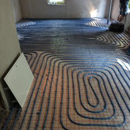 Technika sanitarna Tomasz Bereza - Układanie niebieskich rur ogrzewania podłogowego na folii izolacyjnej z czerwoną siatką w nowym domu w Katowicach, widoczna skrzynka rozdzielcza i fragment osoby przy oknie.