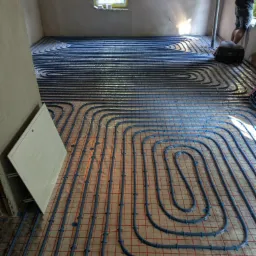 Układanie niebieskich rur ogrzewania podłogowego na folii izolacyjnej z czerwoną siatką w nowym domu w Katowicach, widoczna skrzynka rozdzielcza i fragment osoby przy oknie.