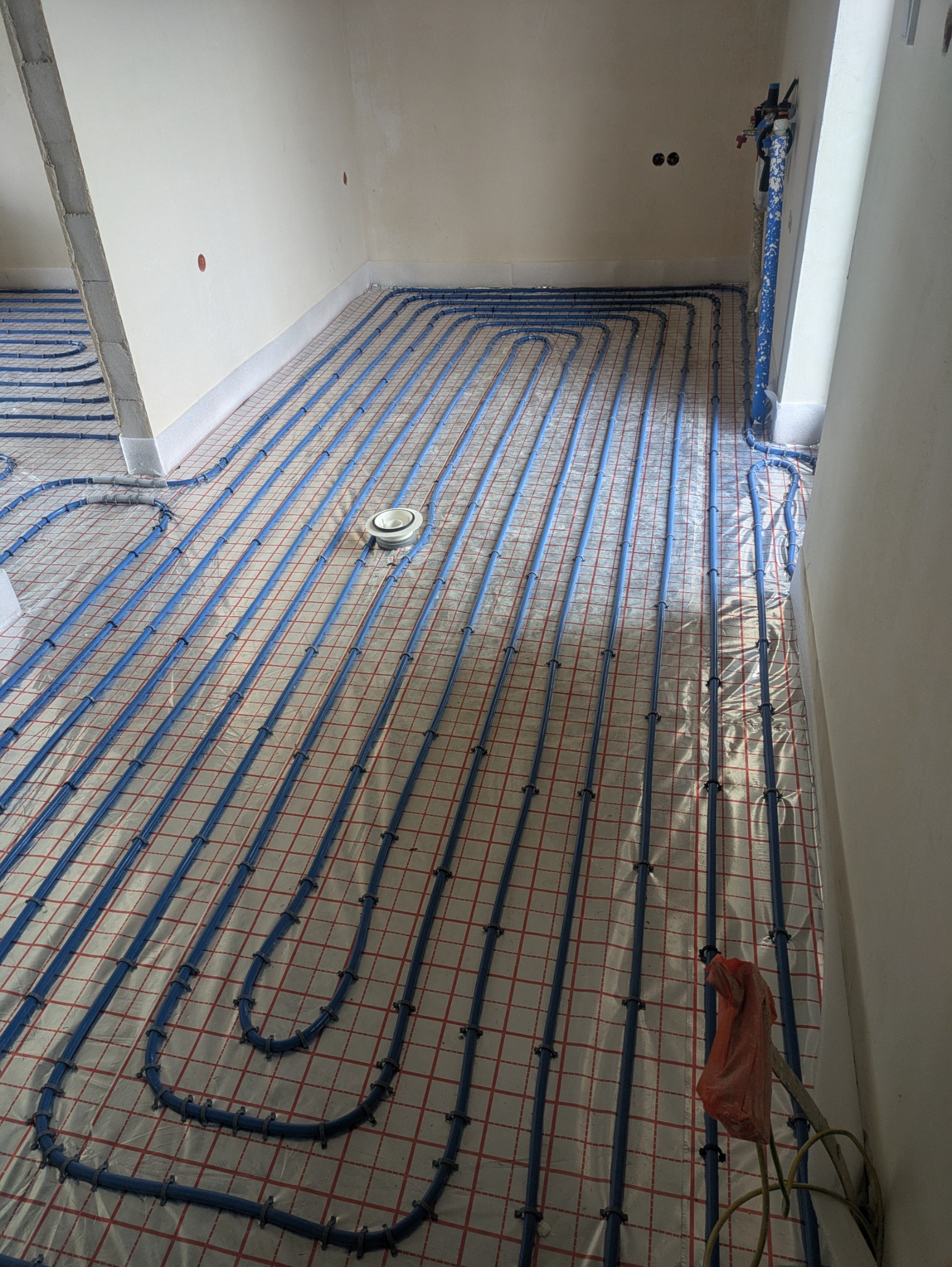 Niedokończona instalacja ogrzewania podłogowego z niebieskich rur na folii z siatką w nowym budynku, widoczny rozdzielacz i podłączenia.