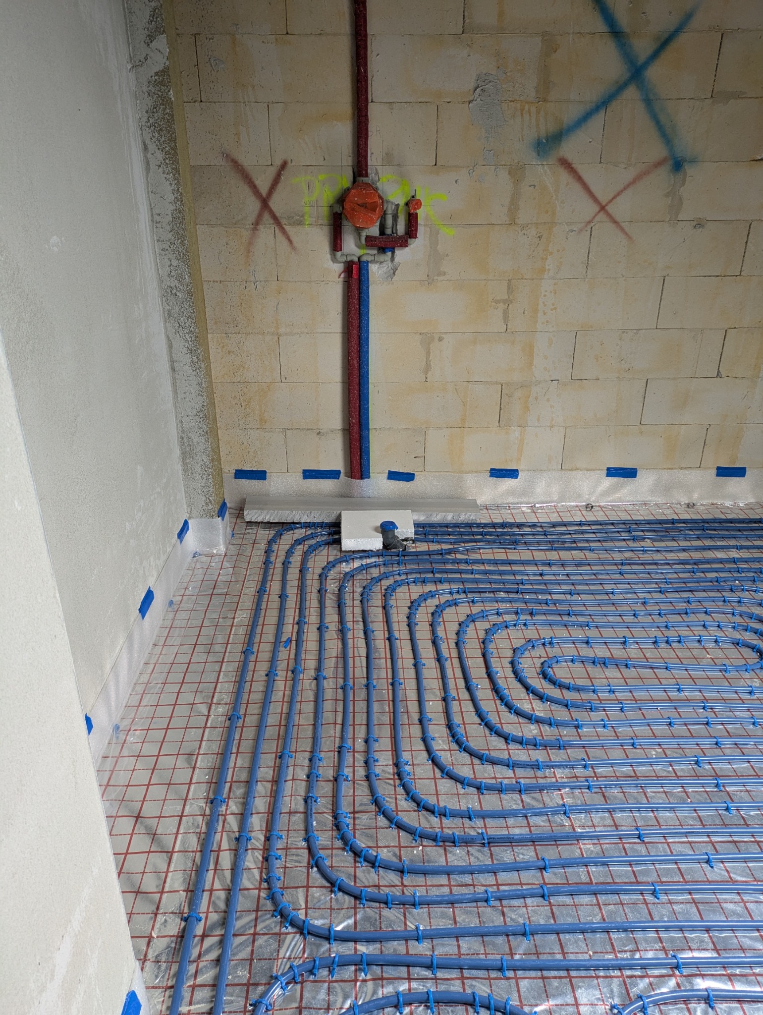 Instalacja ogrzewania podłogowego z niebieskich rur na folii z siatką, widoczne przyłącza do ściany z cegieł betonowych z instalacją wodną w kolorze czerwonym i niebieskim, ściany w surowym stanie.