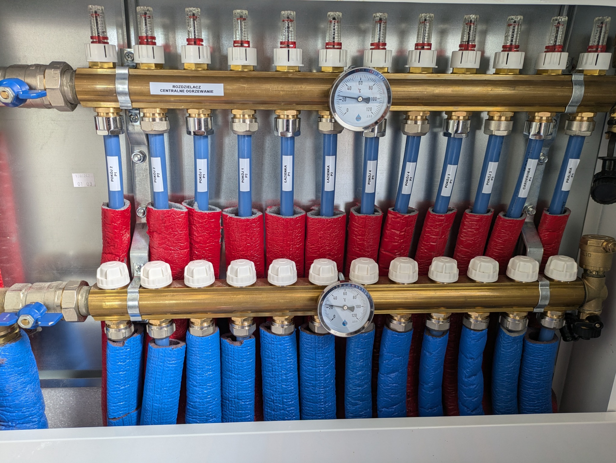 Rozdzielacz centralnego ogrzewania z rurami oznaczonymi dla poszczególnych pomieszczeń, manometry wskazują ciśnienie w instalacji.