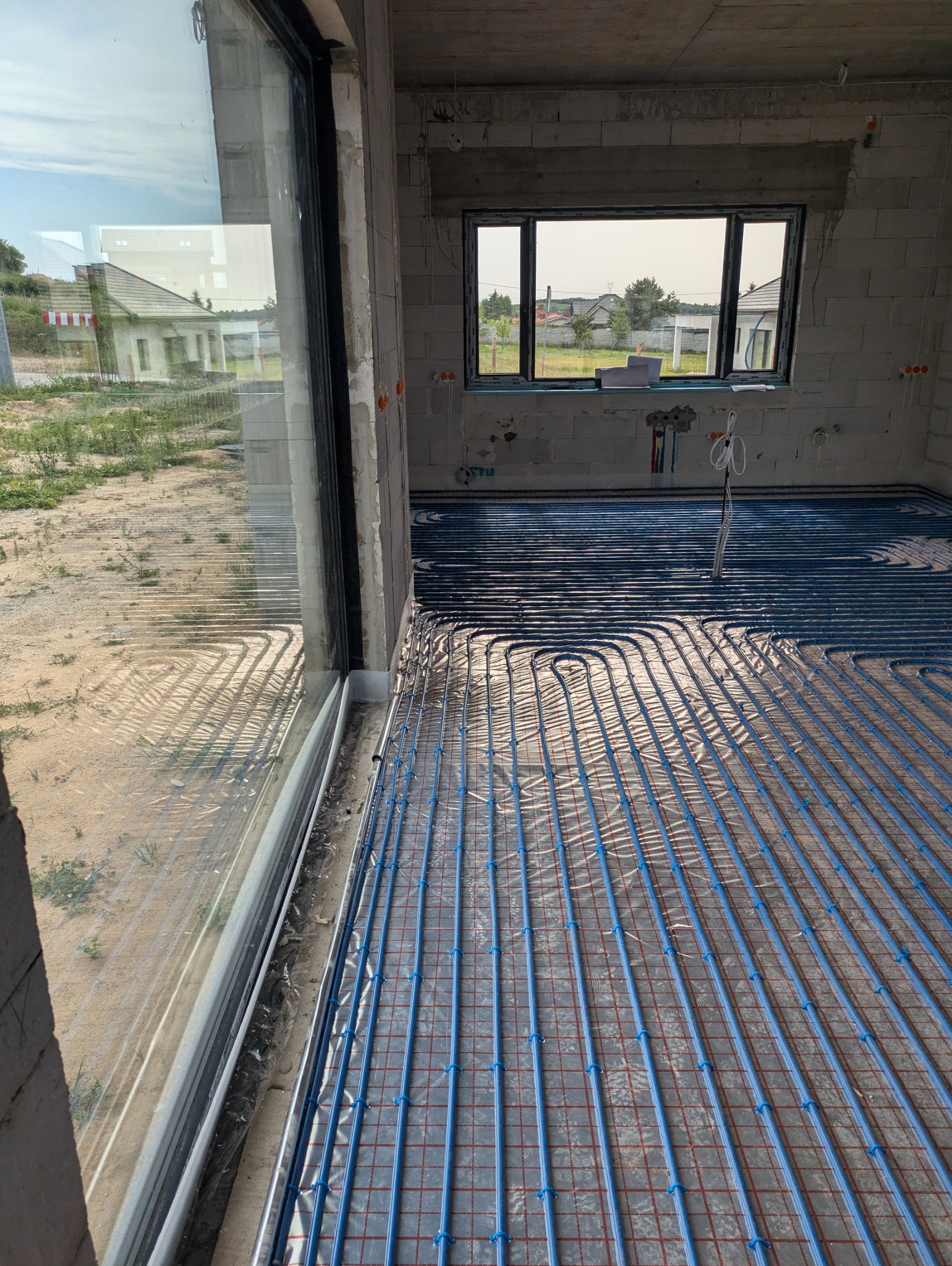 Niedokończona instalacja ogrzewania podłogowego z niebieskich rur na siatce w surowym wnętrzu budynku w trakcie budowy, widok przez duże okno na zewnątrz.