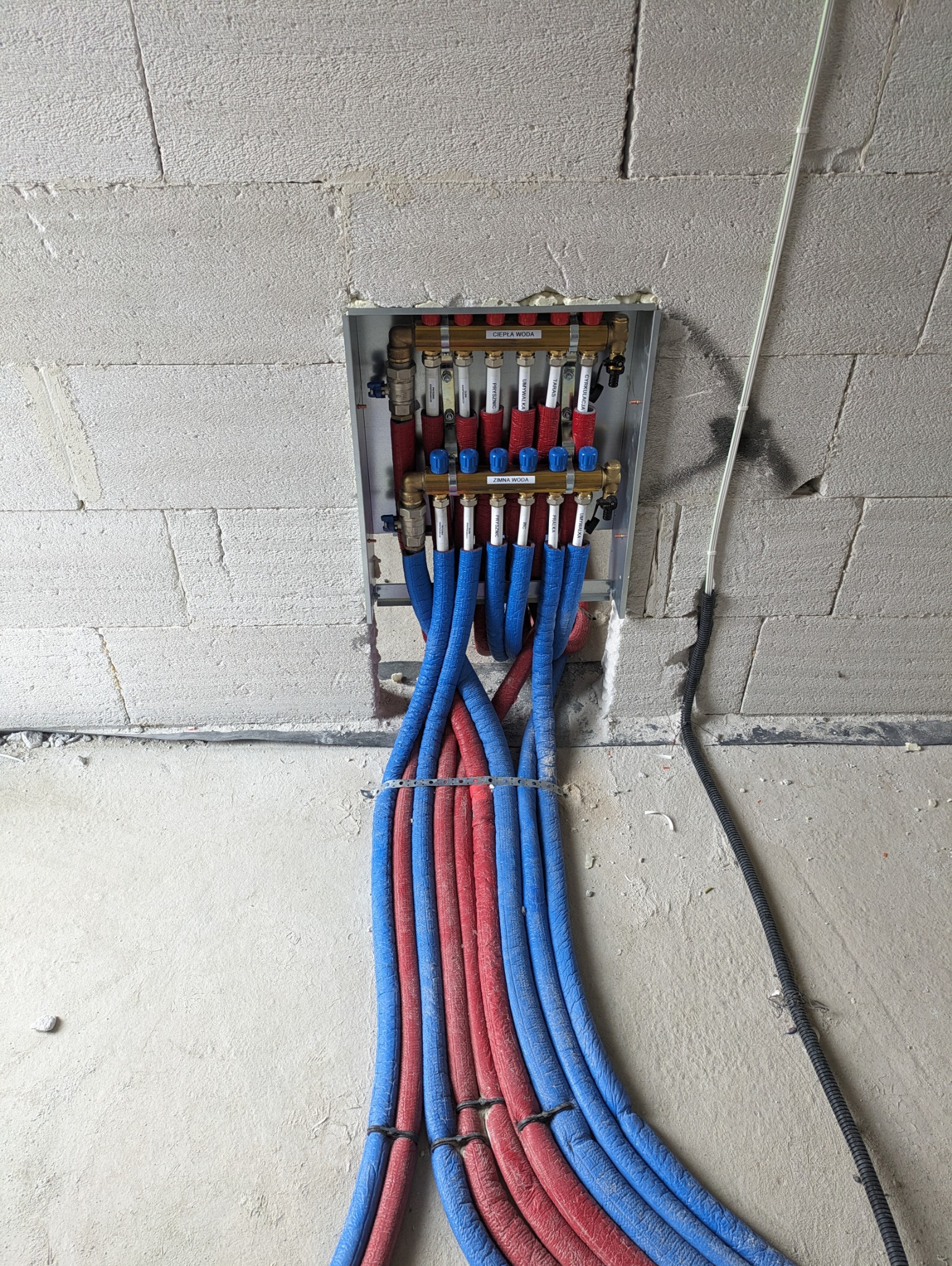 Rozdzielacz instalacji centralnego ogrzewania z podłączonymi, izolowanymi rurami w kolorach czerwonym i niebieskim, zamontowany na surowej ścianie z betonu komórkowego.