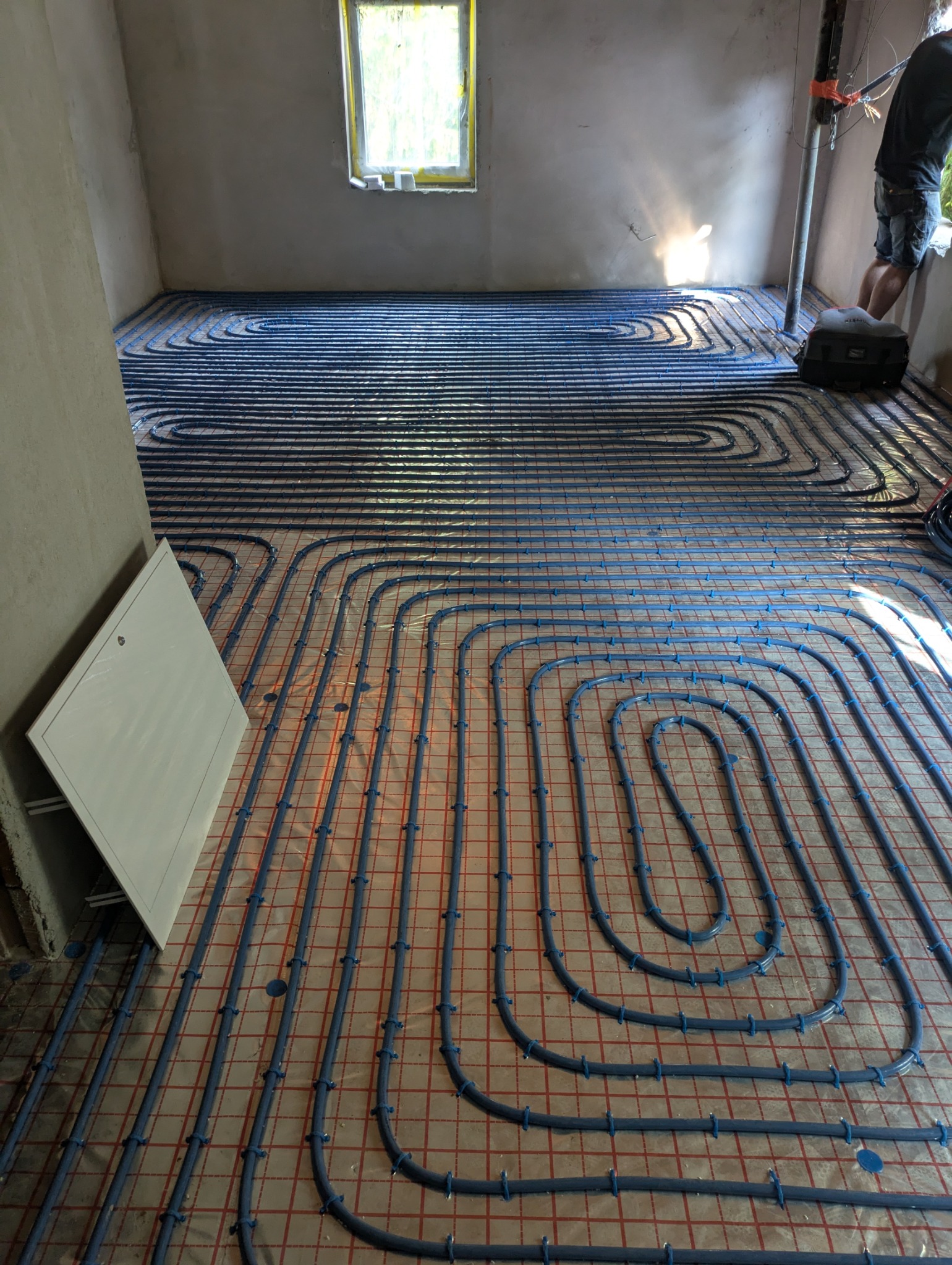 Układanie niebieskich rur ogrzewania podłogowego na folii izolacyjnej z czerwoną siatką w nowym domu w Katowicach, widoczna skrzynka rozdzielcza i fragment osoby przy oknie.