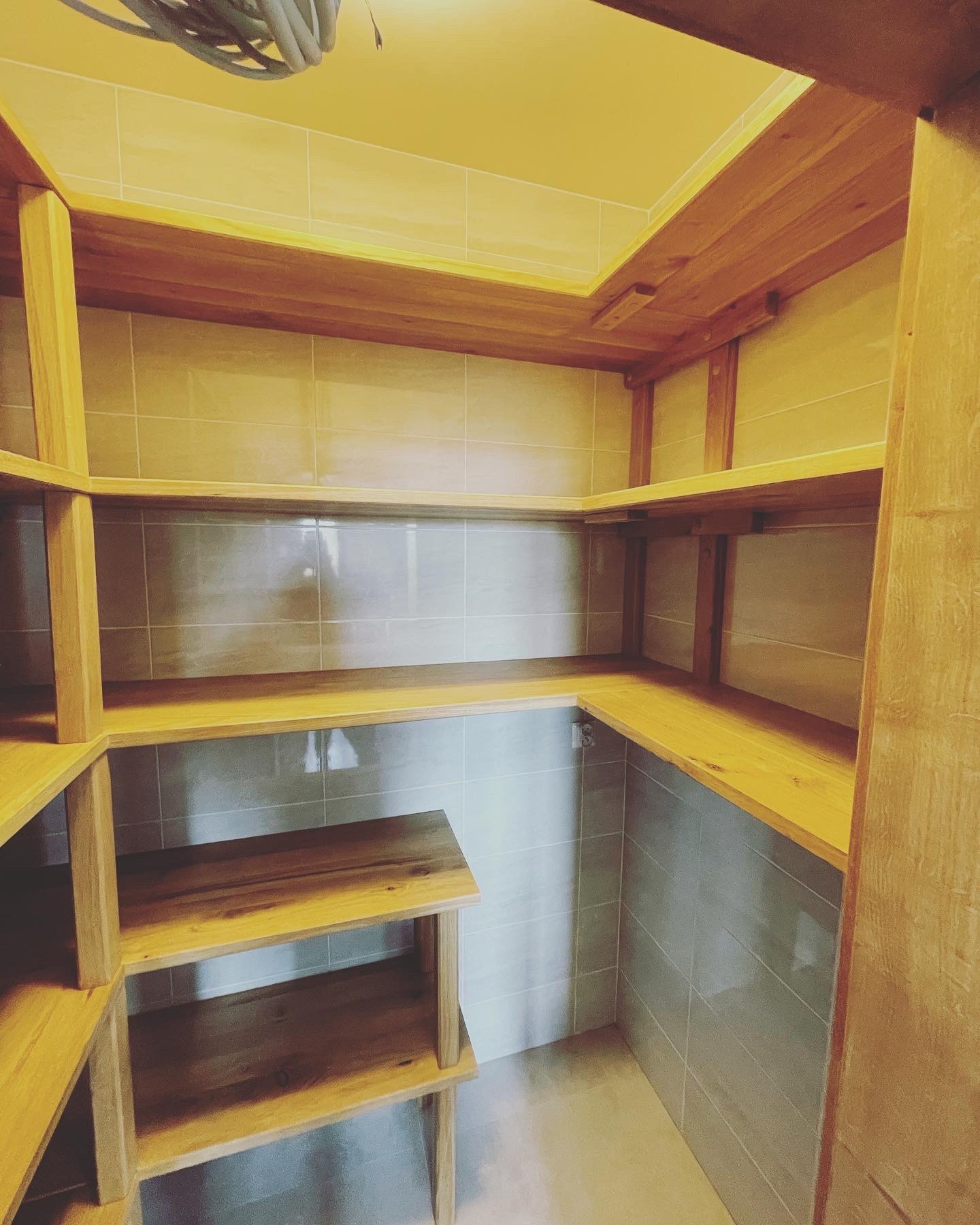 Wnętrze spiżarni z drewnianymi półkami na wymiar, wykończone jasnymi płytkami ceramicznymi, widok z dołu ukazujący konstrukcję narożną i oświetlenie sufitowe.