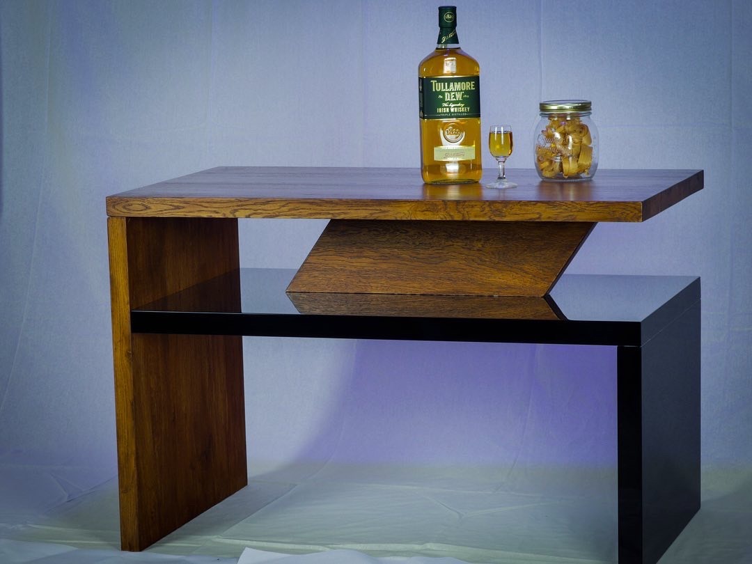 Stolik kawowy z drewna i czarnego szkła, o nowoczesnym designie, z butelką whisky i słoikiem ciasteczek na blacie, oświetlony od dołu niebieskim światłem.