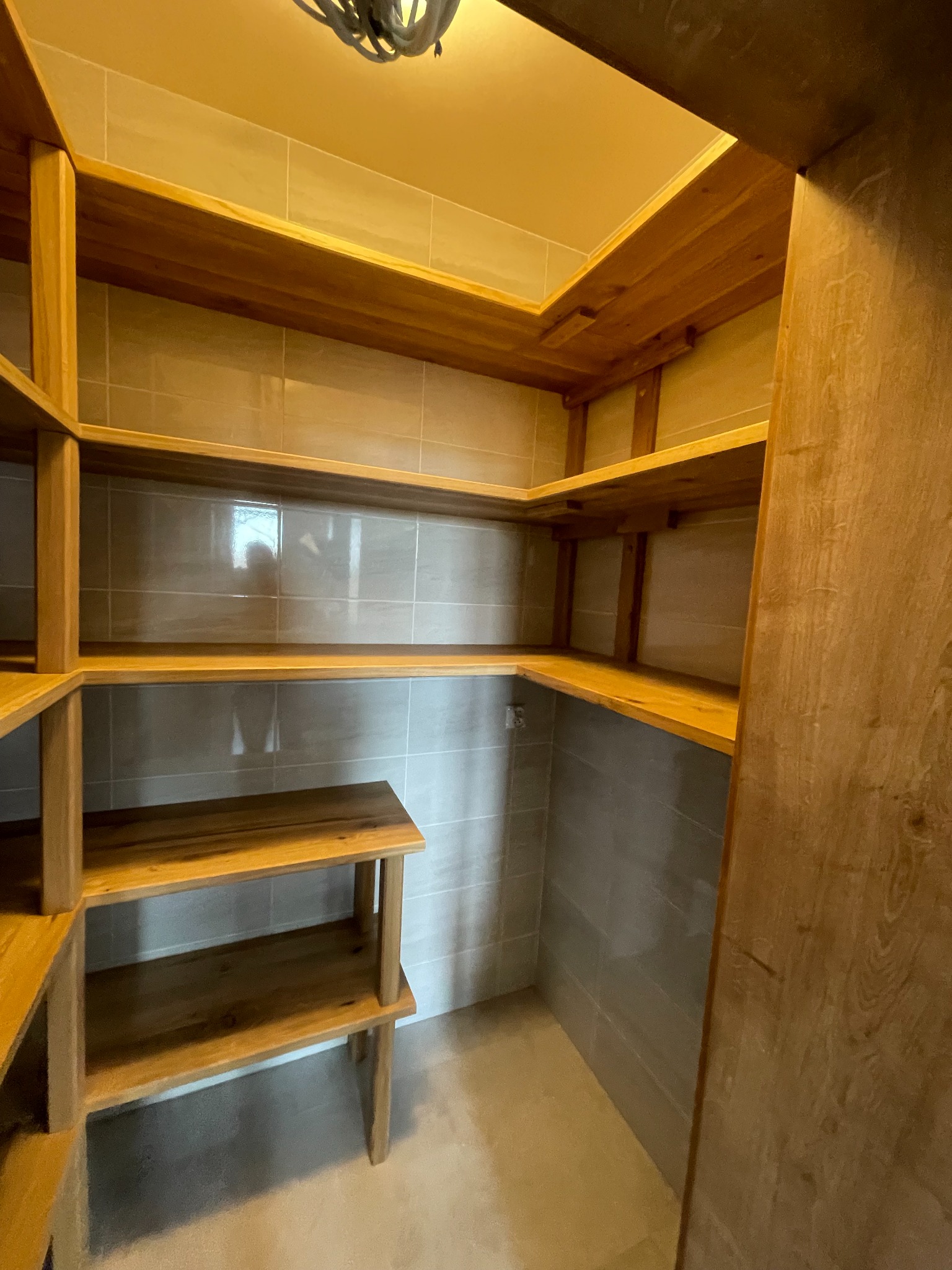 Drewniane półki spiżarni na wymiar z szarymi płytkami na ścianach, oświetlone górnym światłem.