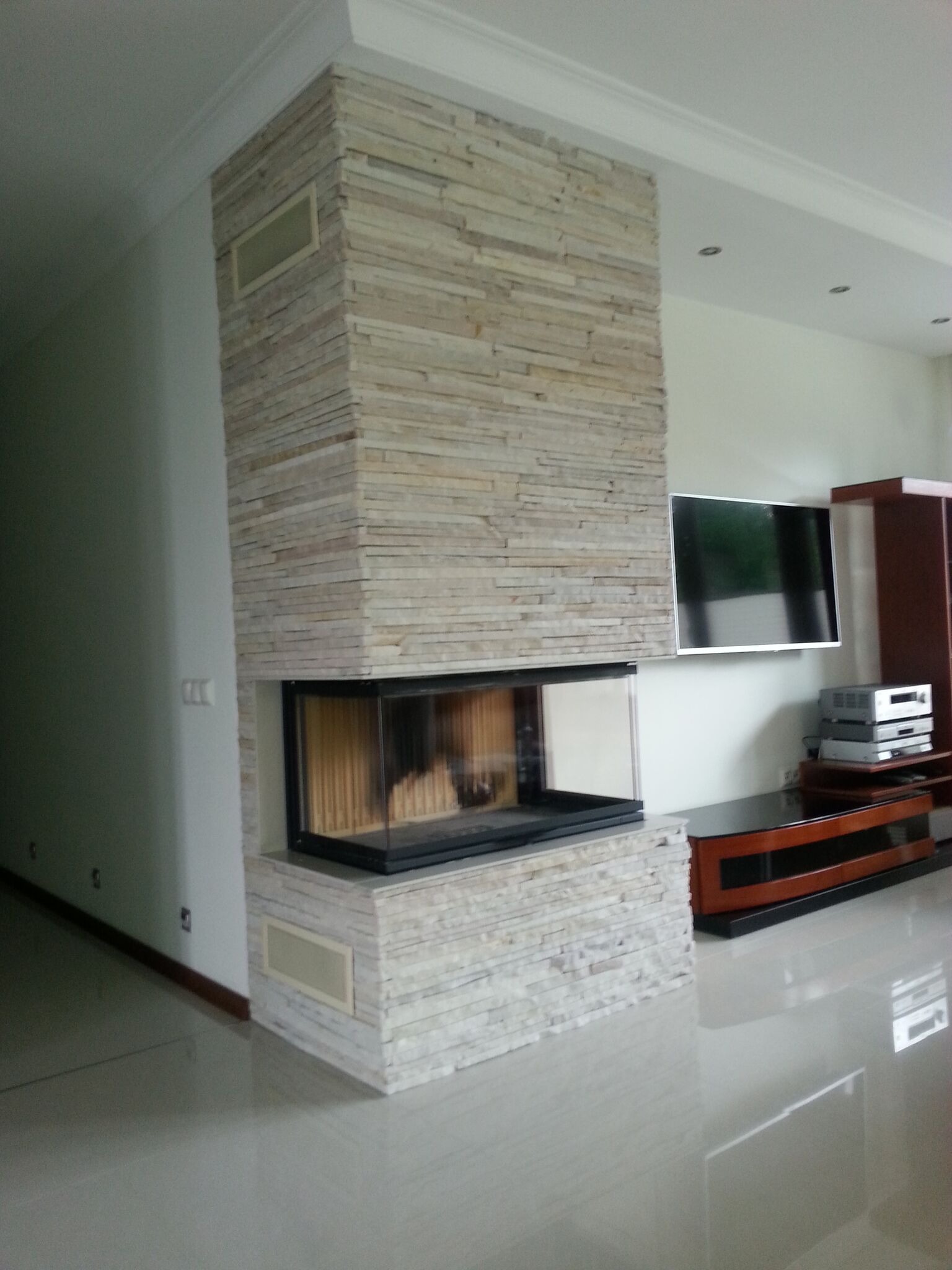 Nowoczesny kominek narożny obłożony kamieniem dekoracyjnym, z trzema szybami, w jasnym salonie z telewizorem na ścianie.