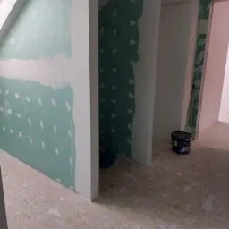Poddasze w trakcie prac wykończeniowych: ściany z płyt kartonowo-gipsowych z widocznymi łączeniami, gruntowane na zielono, z częściowo nałożoną białą gładzią, surowe podłoże podłogowe i wiadro...