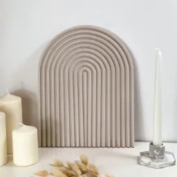 Beżowa, dekoracyjna płyta z motywem łuków, ustawiona obok białych świec o różnej wysokości i suszonych traw na jasnym blacie.