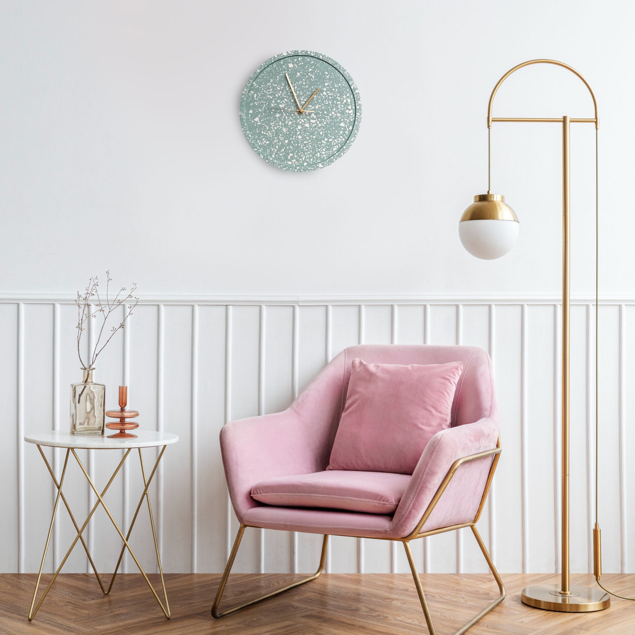 Nowoczesny salon z pudrowo-różowym fotelem na złotych nóżkach, marmurowym stolikiem z dekoracjami i złotą lampą łukową na tle białej ściany z panelami i zegarem w stylu lastryko.
