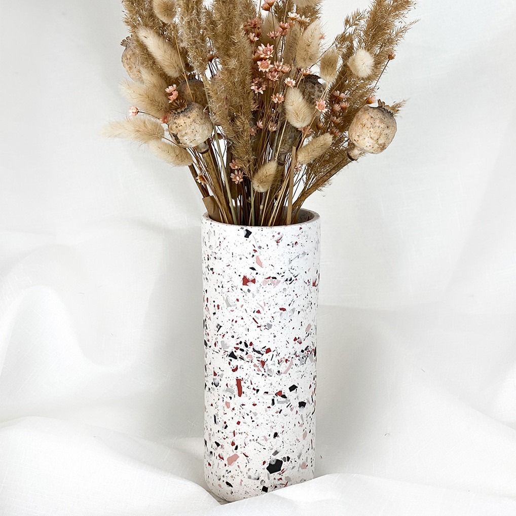 Wysoki, cylindryczny wazon z terazzo w kolorze białym, wypełniony suszonymi trawami, makówkami i drobnymi różowymi kwiatami na tle białej, pofałdowanej tkaniny.