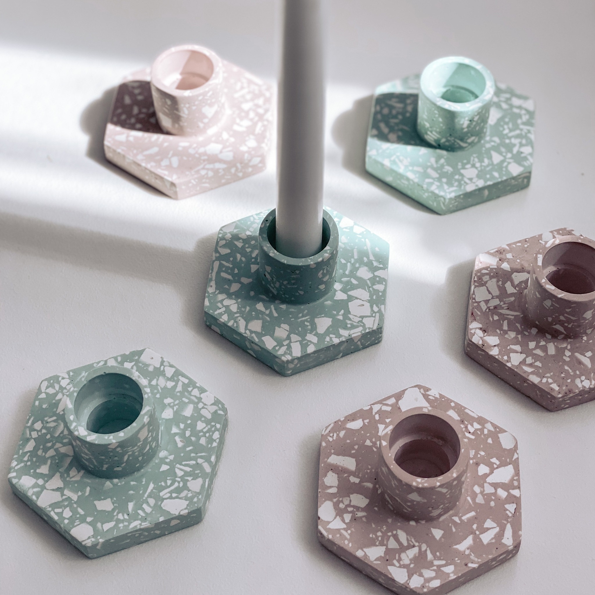 Pięć betonowych świeczników w pastelowych kolorach (różowy, miętowy) o fakturze lastryko, ułożonych na białym tle w jasnym świetle, jeden z białą świecą.