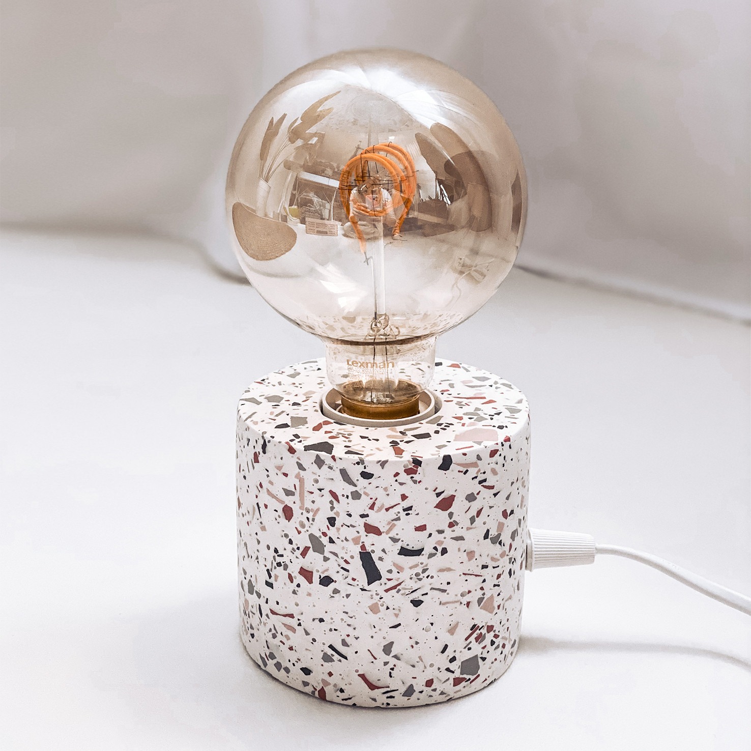 Lampa stołowa z okrągłą żarówką i podstawą z lastryko, widoczny kabel zasilający.