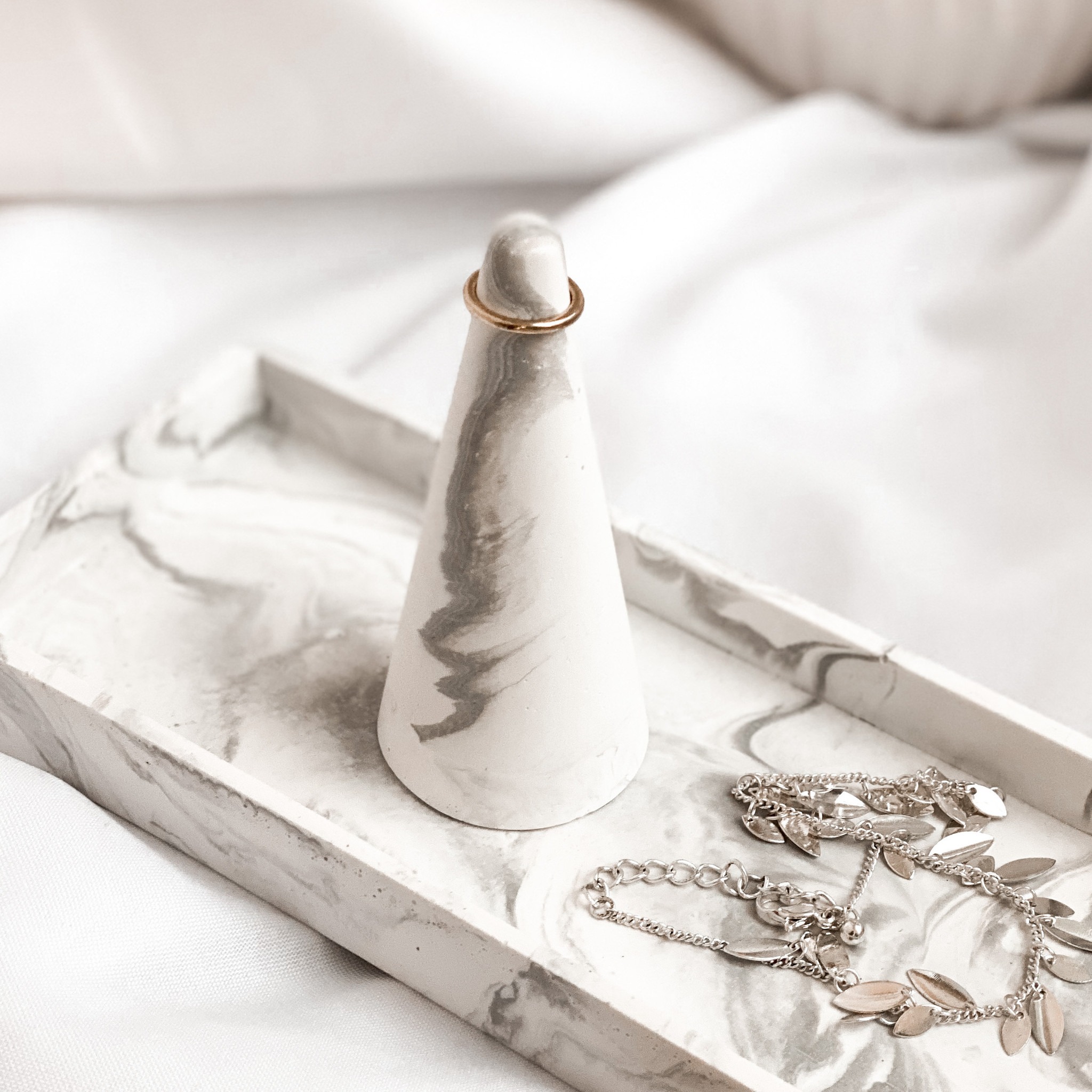 Betonowa podstawka na biżuterię z marmurkowym wzorem, złoty pierścionek i srebrny łańcuszek z listkami na tacy, miękkie, rozproszone światło.