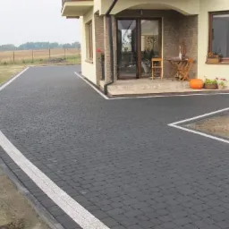 Czarna kostka brukowa ułożona przed domem z białymi akcentami, rabata z ziemią, dekoracje przed wejściem.