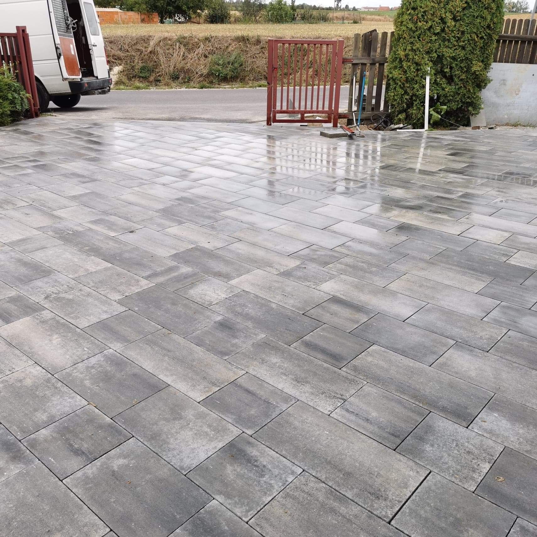 Świeżo ułożona, mokra kostka brukowa w odcieniach szarości, ułożona we wzór prostokątów, z widoczną bramką i samochodem dostawczym w tle.