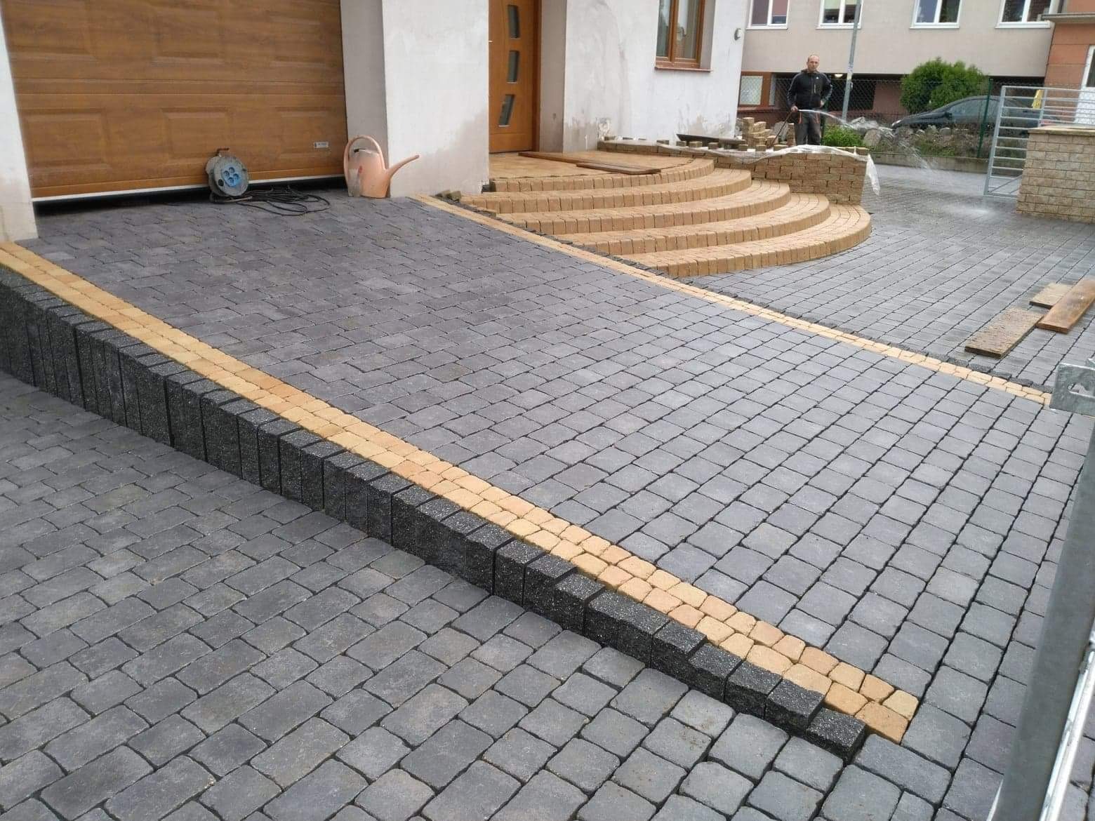 Ukończony podjazd z kostki brukowej w odcieniach szarości i beżu, z widocznymi schodami wejściowymi i osobą podlewającą rośliny w tle.