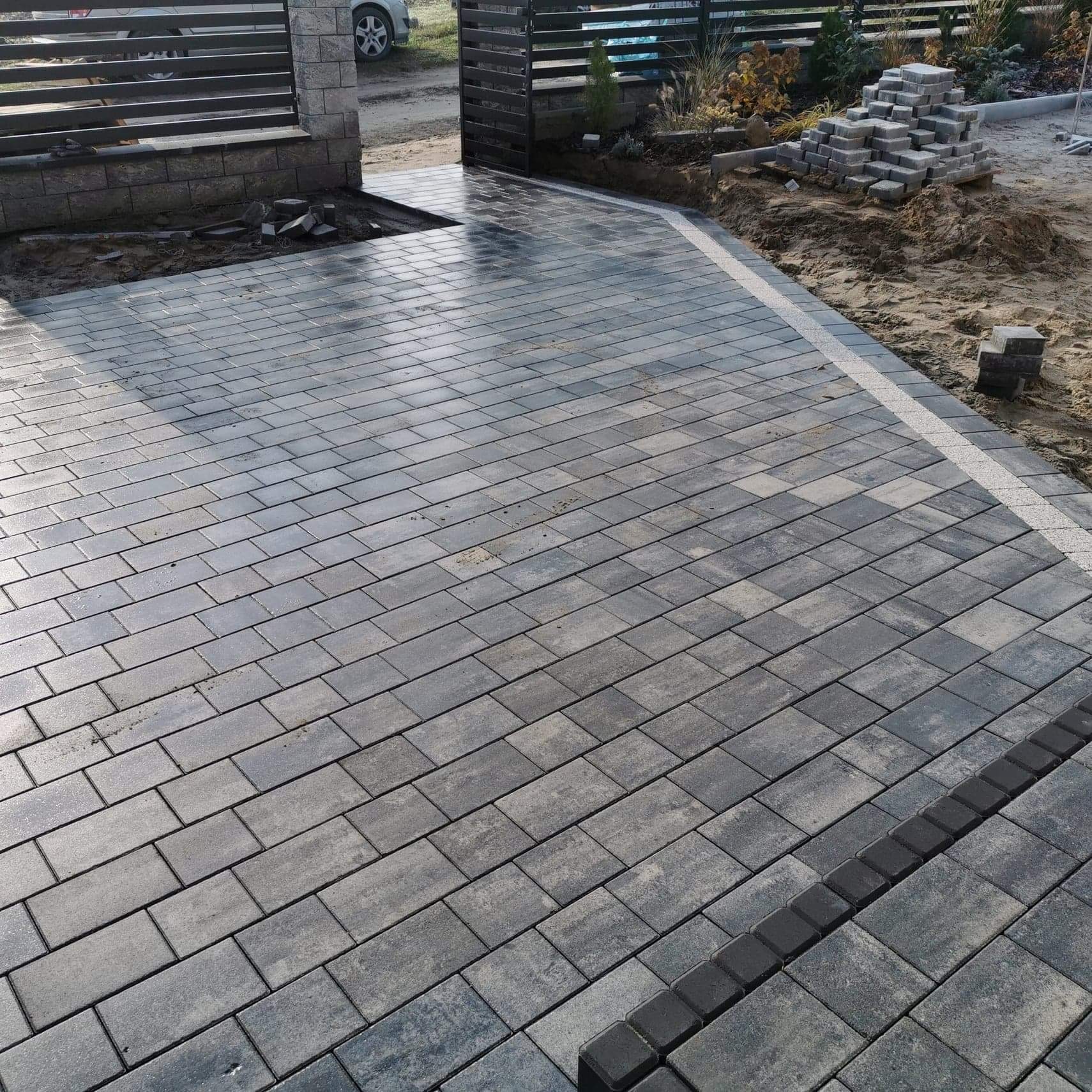 Ułożona szara kostka brukowa tworząca podjazd, z widocznym obramowaniem z ciemniejszych kostek i stertą kostki przygotowanej do dalszej pracy, na tle ogrodzenia i częściowo widocznego samochodu.