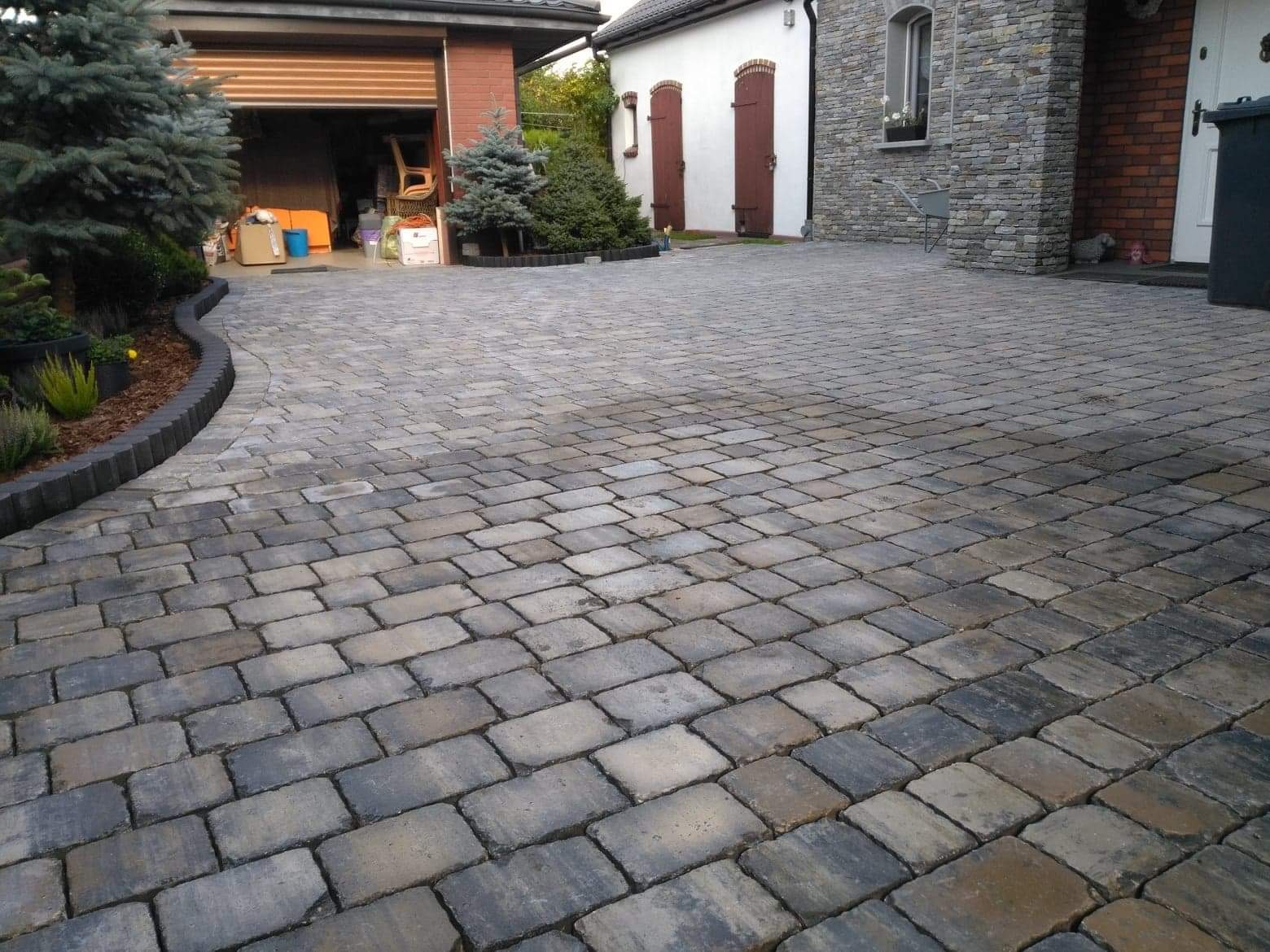 Szeroki, wyłożony kostką brukową podjazd do domu z garażem, z obrzeżem z palisady i roślinnością, widok na budynek z kamienną elewacją i brązowymi okiennicami.