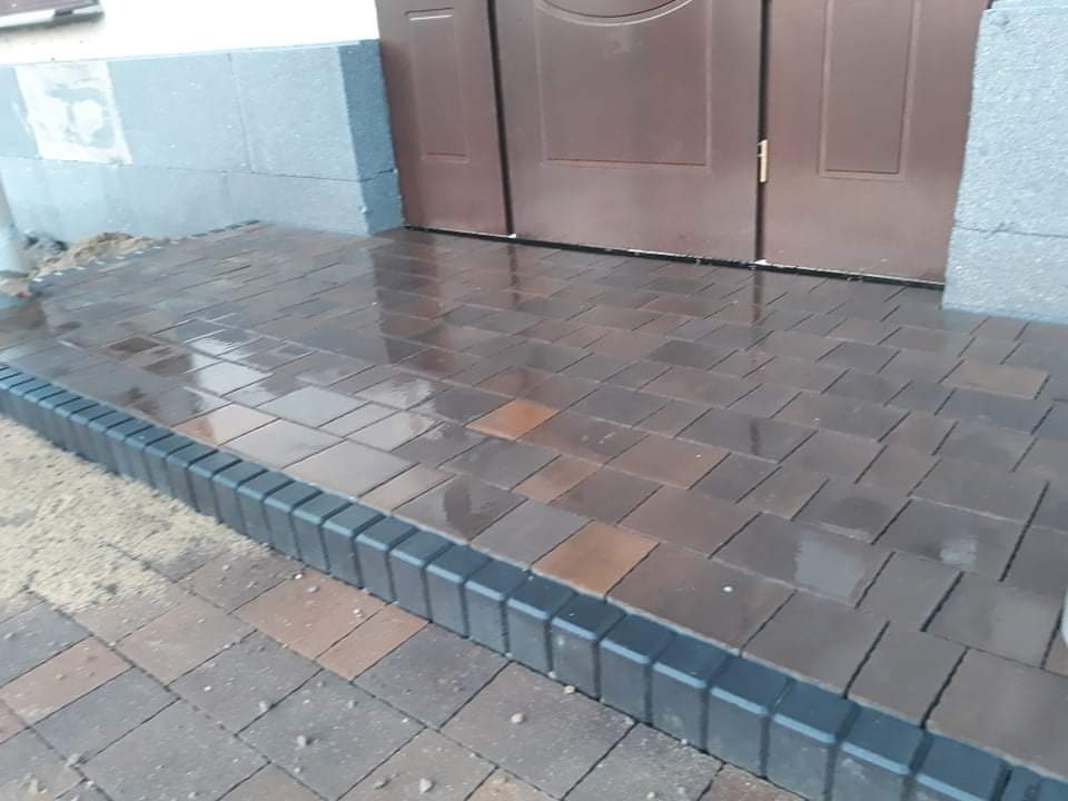 Świeżo ułożona, mokra kostka brukowa w odcieniach brązu i szarości prowadząca do wejścia z brązowymi drzwiami, obrzeże z ciemnej kostki, widoczne ślady piasku.