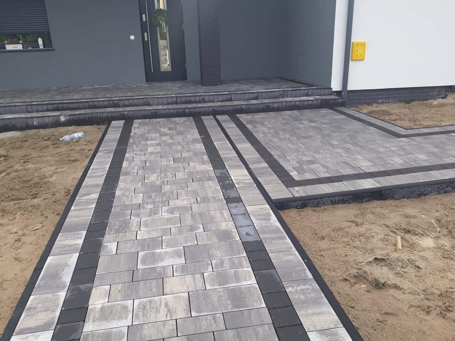 Ułożona kostka brukowa w odcieniach szarości i czerni prowadząca do wejścia domu z widoczną butelką wody na piasku obok.