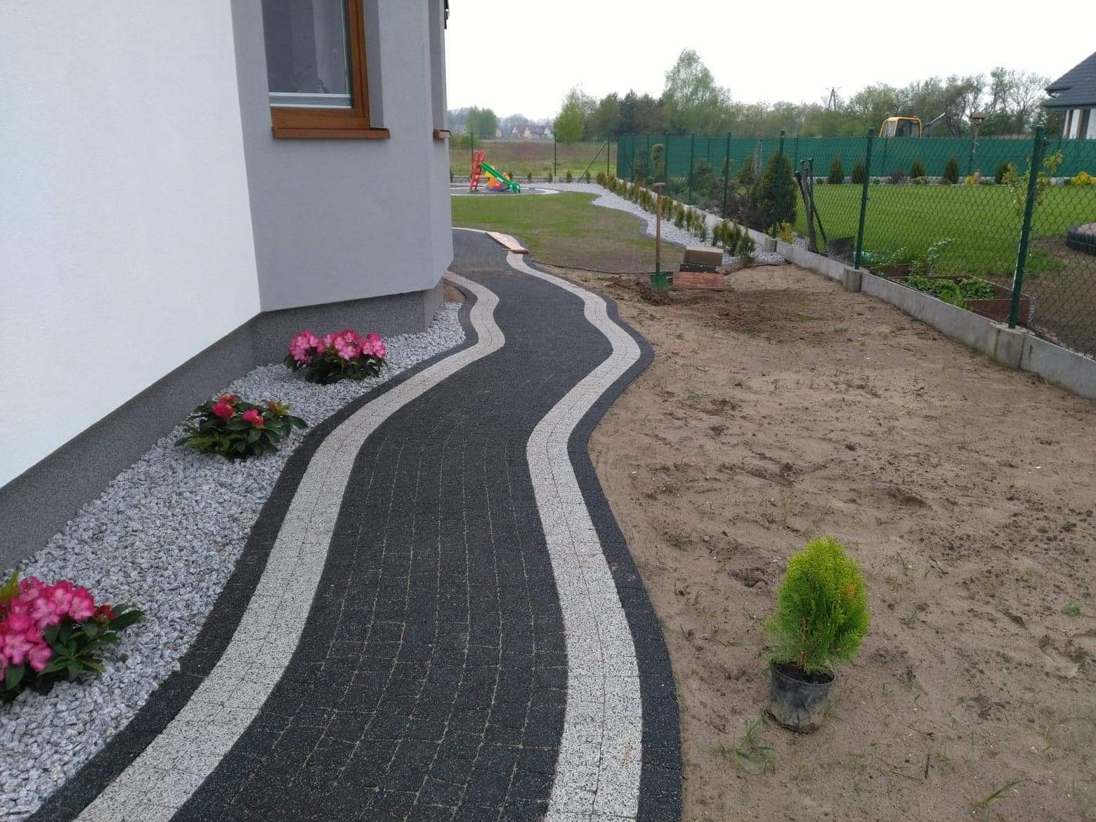 Kręta ścieżka z ciemnej kostki brukowej z jasnymi pasami obrzeży, obsadzona różowymi kwiatami i szarym żwirem przy ścianie domu, w tle trawnik z placem zabaw i ogrodzeniem.