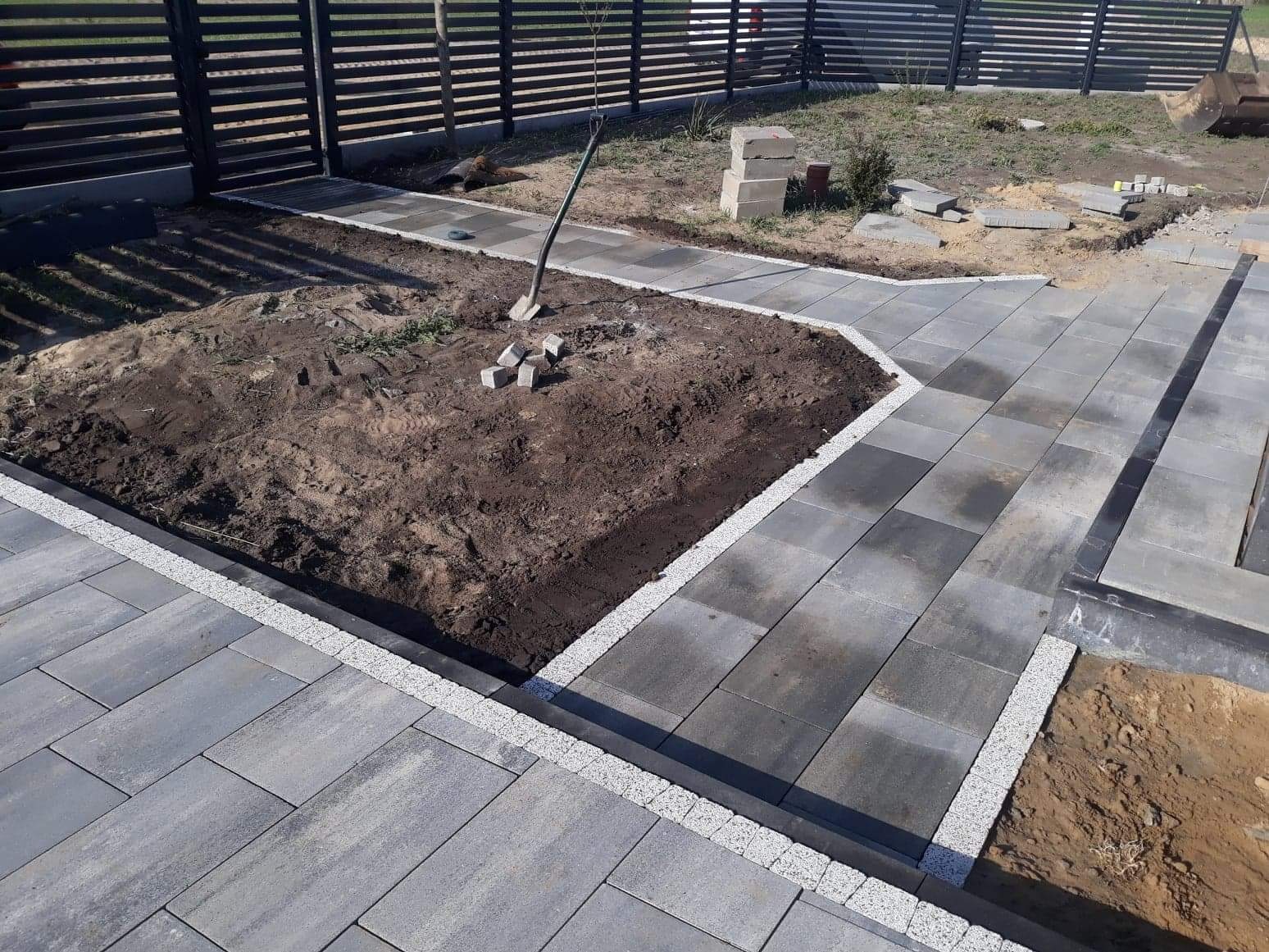 Fragment ścieżki z szarej kostki brukowej, obrzeże z jasnego kruszywa, obok rabata z ziemią i pojedyncza łopata, w tle metalowe ogrodzenie.