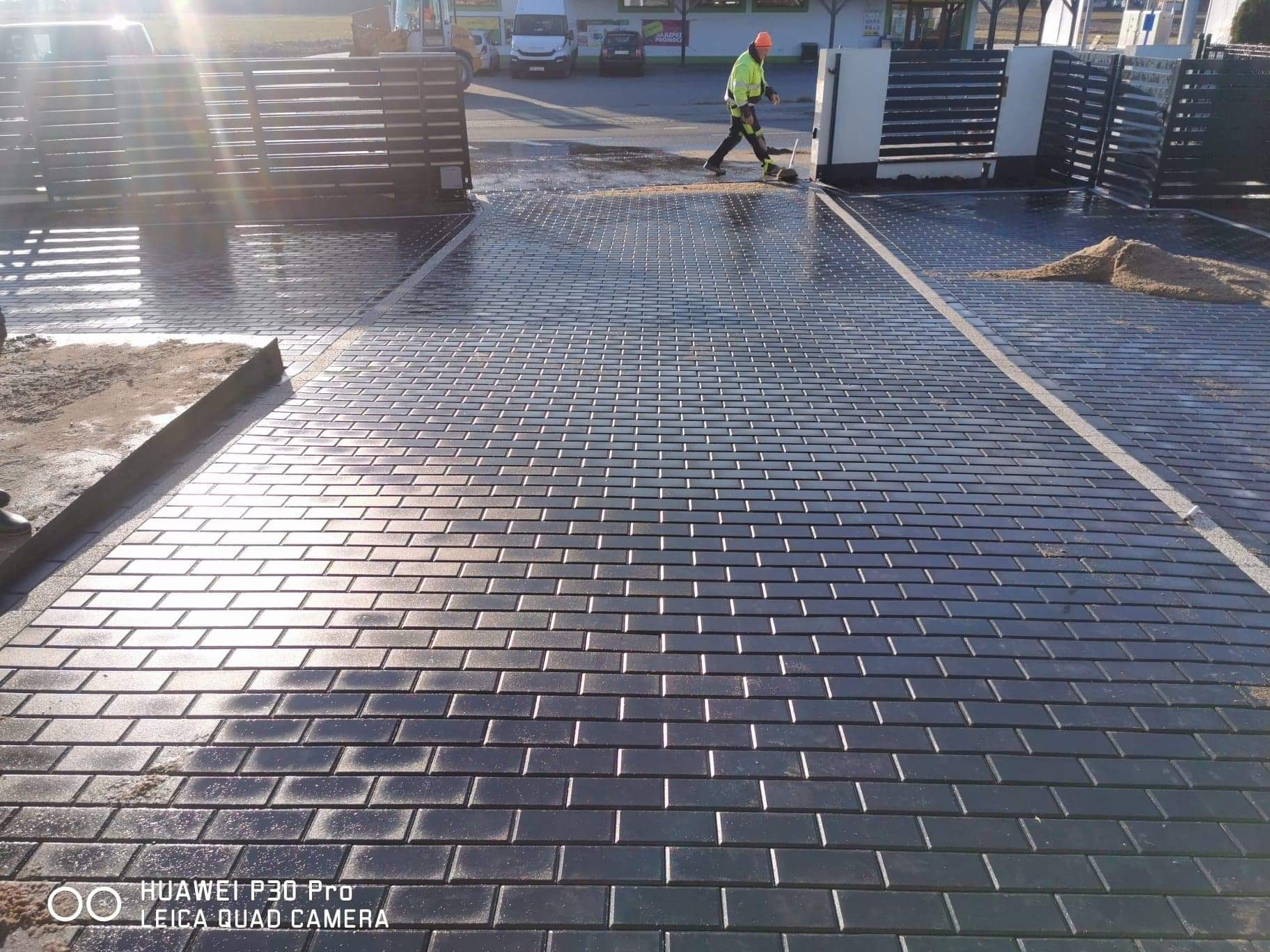 Świeżo ułożona, czarna kostka brukowa w kształcie cegiełek, mokra od wody, z widoczną linią dylatacyjną, pracownik w odblaskowej kamizelce zamiata resztki piasku, brama w tle.