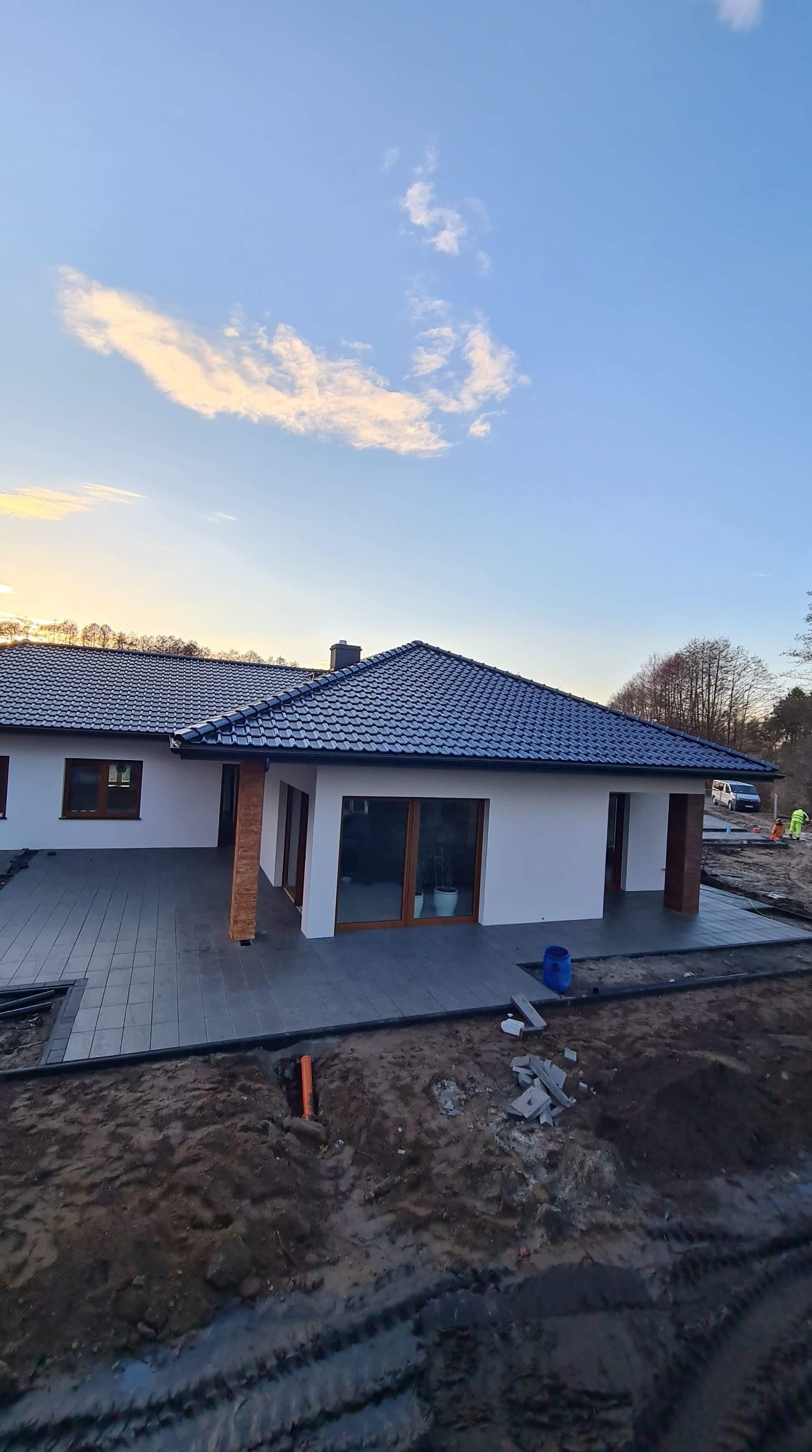 Nowoczesny dom z ciemną dachówką i szarym tarasem z kostki, widok z góry na częściowo zagospodarowany teren, rura w ziemi, w tle pracownicy w odblaskowych kamizelkach.