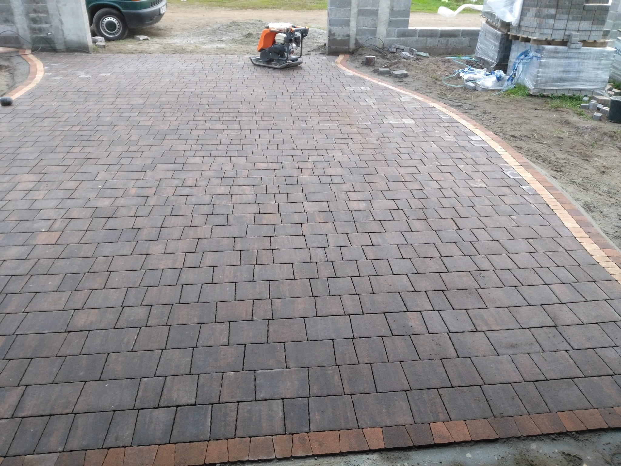 Układanie ciemnobrązowej kostki brukowej z obramowaniem z czerwonej kostki, zagęszczarka wibracyjna na powierzchni, w tle palety z kostką i budowa ogrodzenia.
