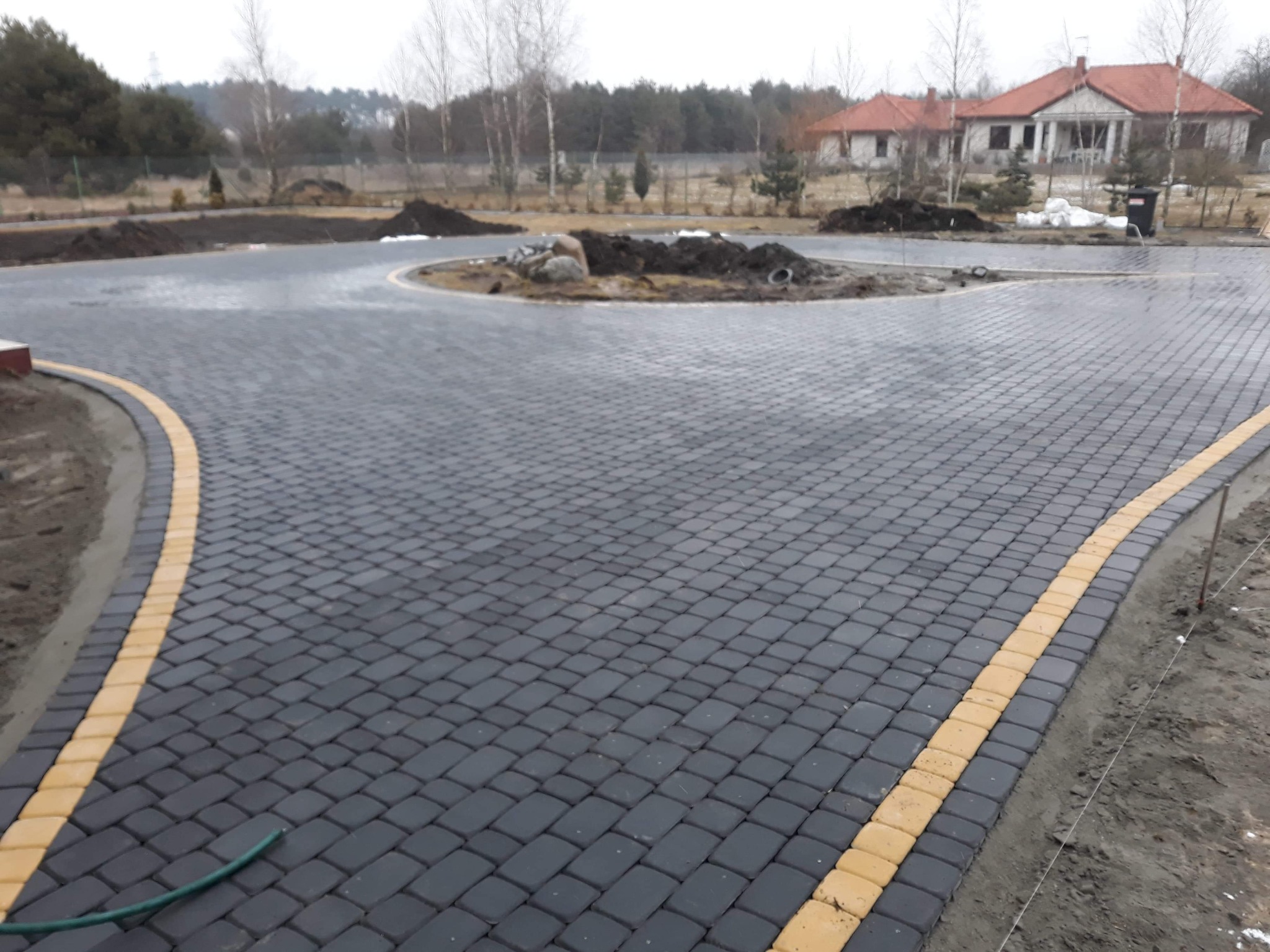 Nowo ułożona, ciemnoszara kostka brukowa z żółtymi obrzeżami tworząca rozwidlenie drogi i okrągły klomb z ziemią i kamieniami na posesji z domem w tle.