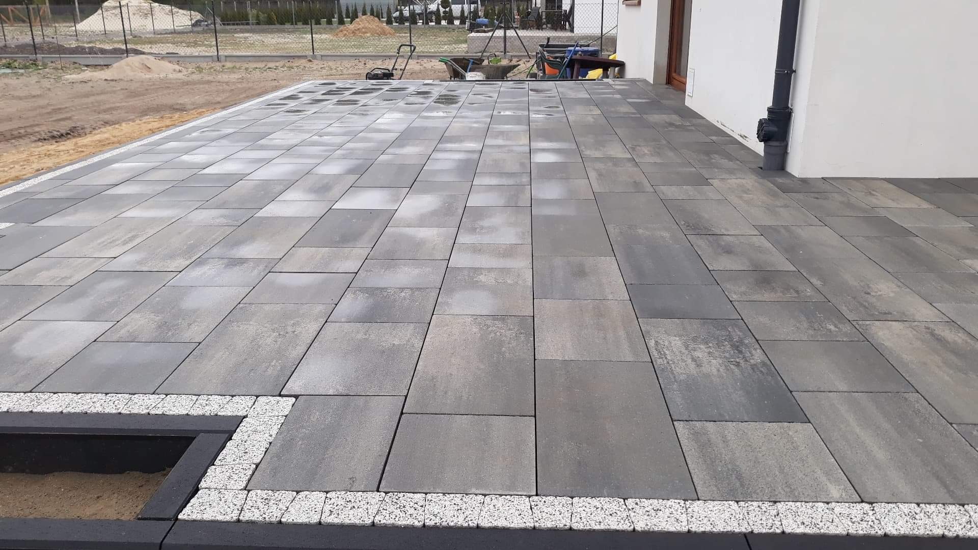 Ułożona, szara kostka brukowa o prostokątnym kształcie, obrzeża wykończone drobnym, białym kamieniem, widoczny fragment rabaty z piaskiem, w tle narzędzia ogrodowe i teren budowy.