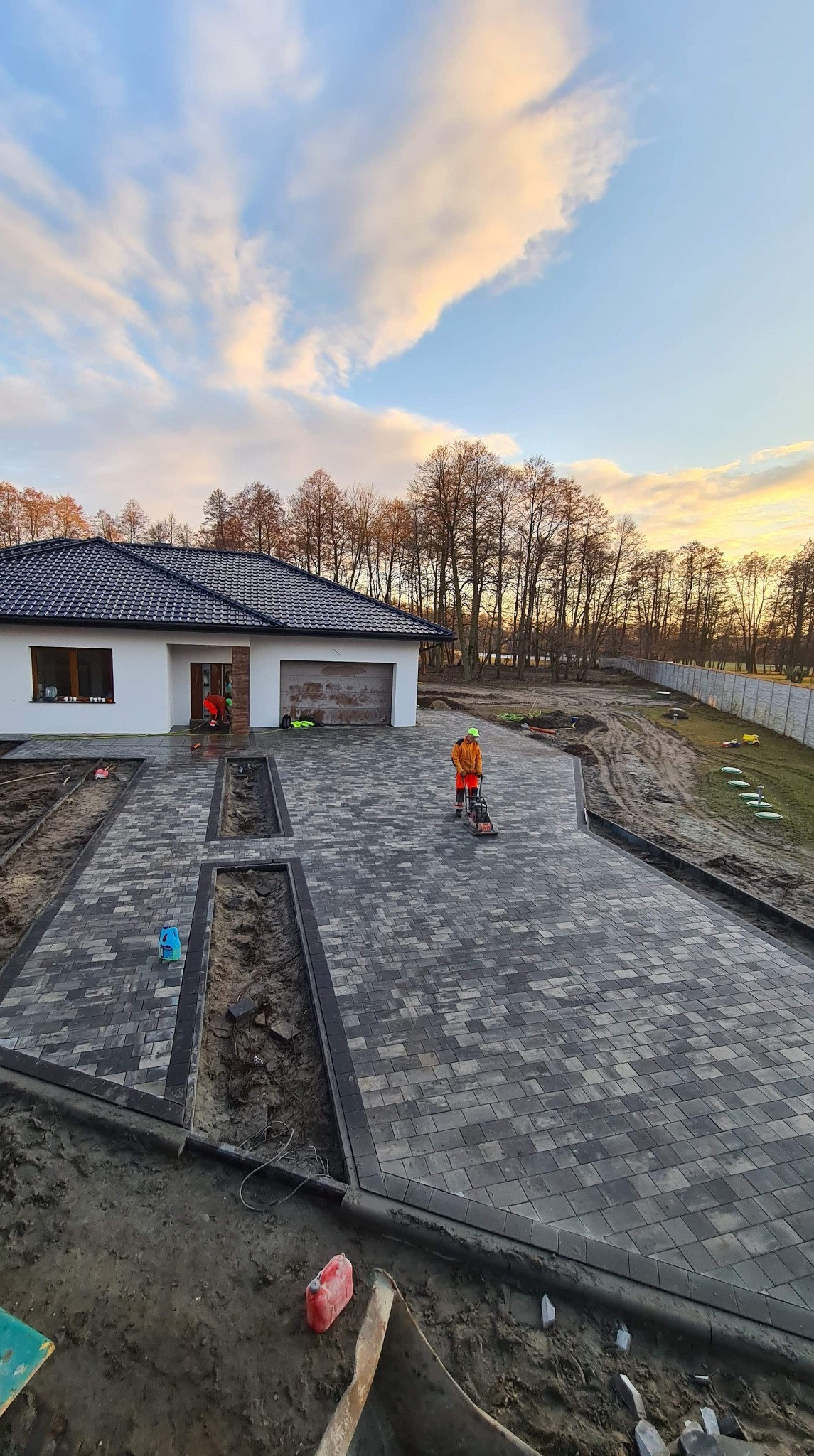 Układanie szarej kostki brukowej na podjeździe przed domem jednorodzinnym; pracownik zagęszcza powierzchnię wibratorem, drugi pracownik czyści kostkę przy wejściu do domu, widoczne przygotowane...