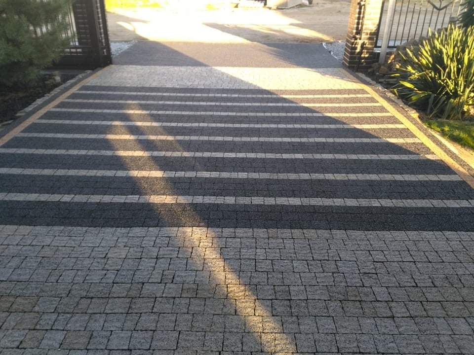 Ułożona nawierzchnia z kostki granitowej w odcieniach szarości, z naprzemiennymi pasami ciemniejszej i jaśniejszej kostki, prowadząca do bramy wjazdowej; widoczny fragment ogrodu po prawej stronie.