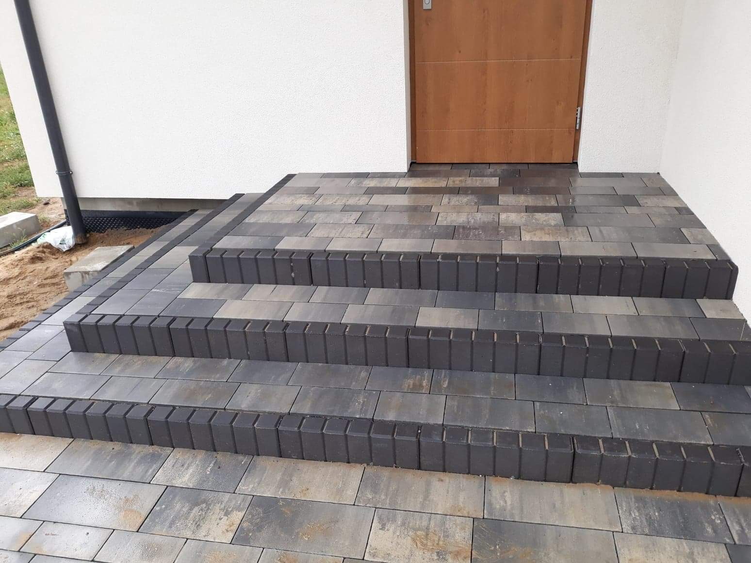 Schody wejściowe do domu wyłożone kostką w odcieniach szarości i grafitu, z widocznym fragmentem elewacji i drewnianych drzwi.