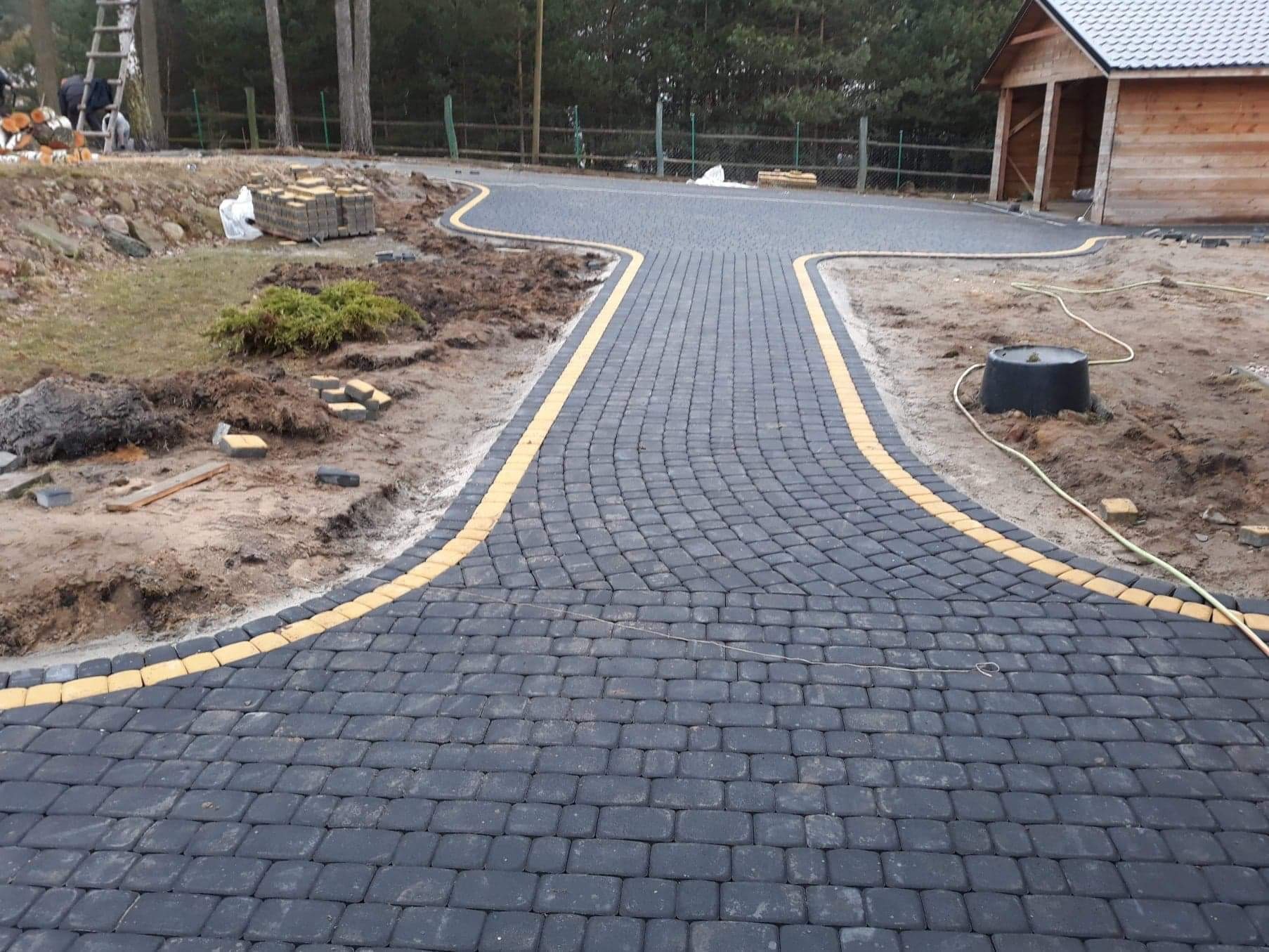 Ułożona czarna kostka brukowa z żółtymi obrzeżami tworząca ścieżkę rozwidlającą się na podjeździe, w tle sterta kostki, drewniana altana i drzewa.