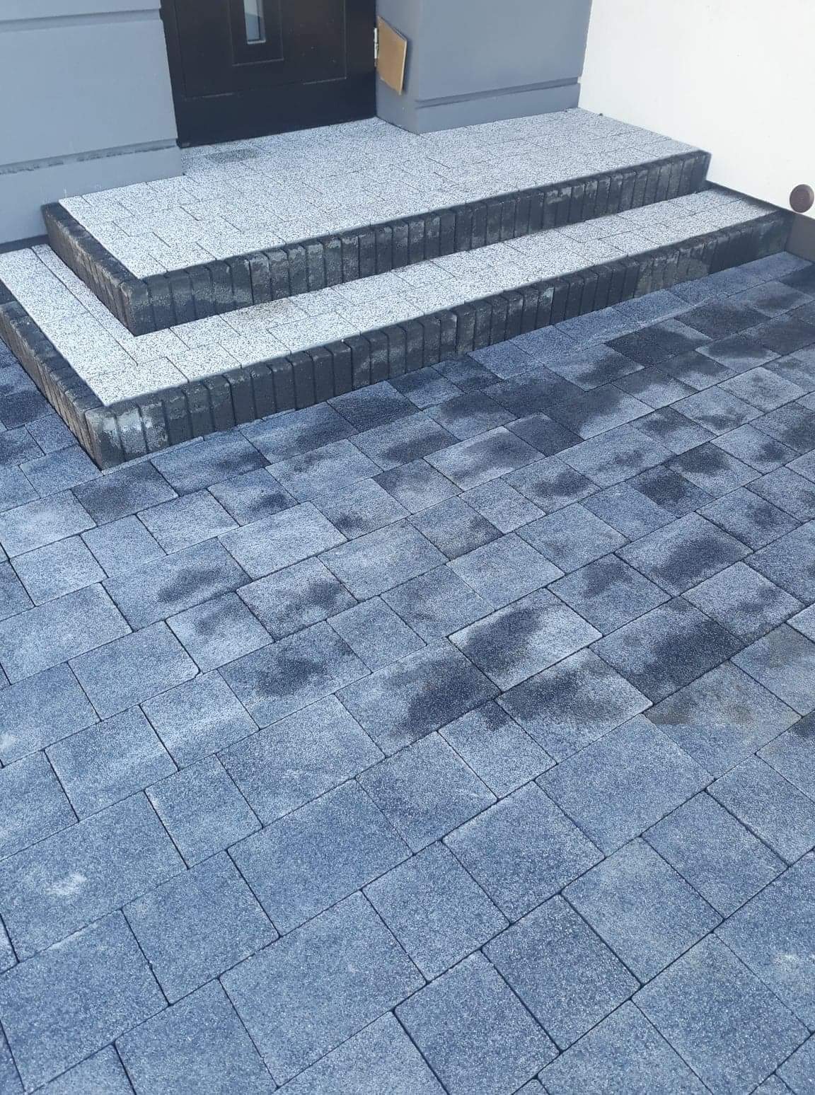 Dwu-stopniowe schody wejściowe wyłożone jasnoszarą kostką z ciemnoszarym obramowaniem, prowadzące do drzwi; obszar przed schodami wyłożony niebiesko-szarą kostką brukową.