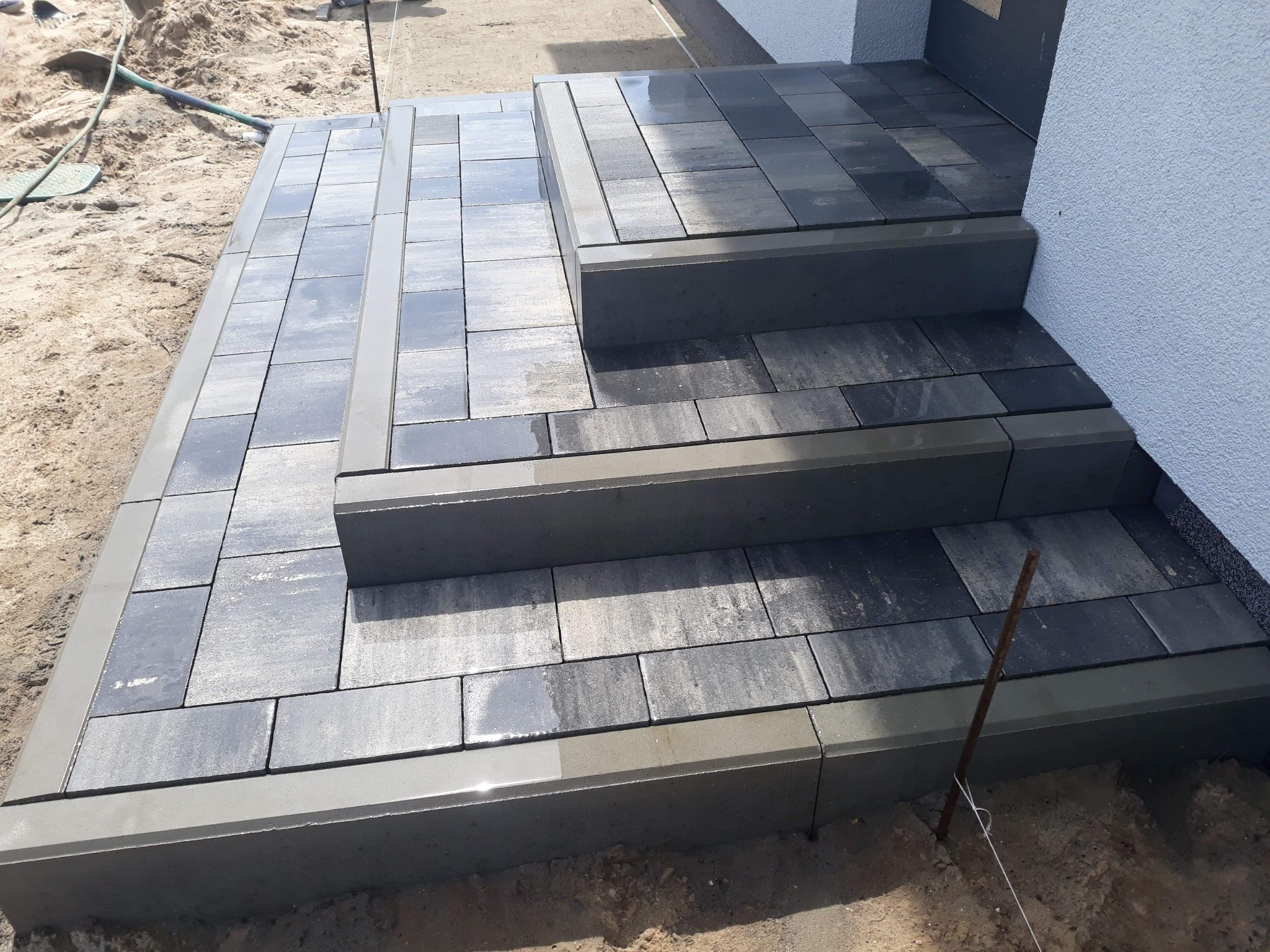 Schody wejściowe do domu wyłożone szarą kostką brukową, widoczne ślady wilgoci na powierzchni, obrzeża z betonu, w tle piasek i fragment elewacji budynku.