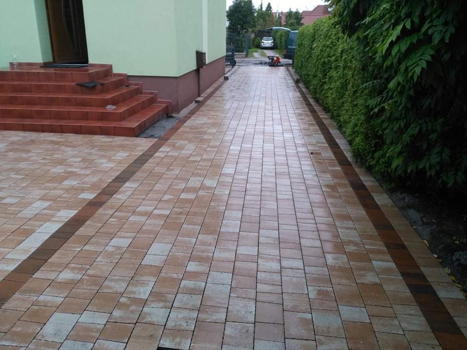 Ułożona kostka brukowa w kolorach beżu i brązu tworzy ścieżkę od schodów domu do ulicy, obok żywopłotu; widoczna maszyna do cięcia kostki w tle.