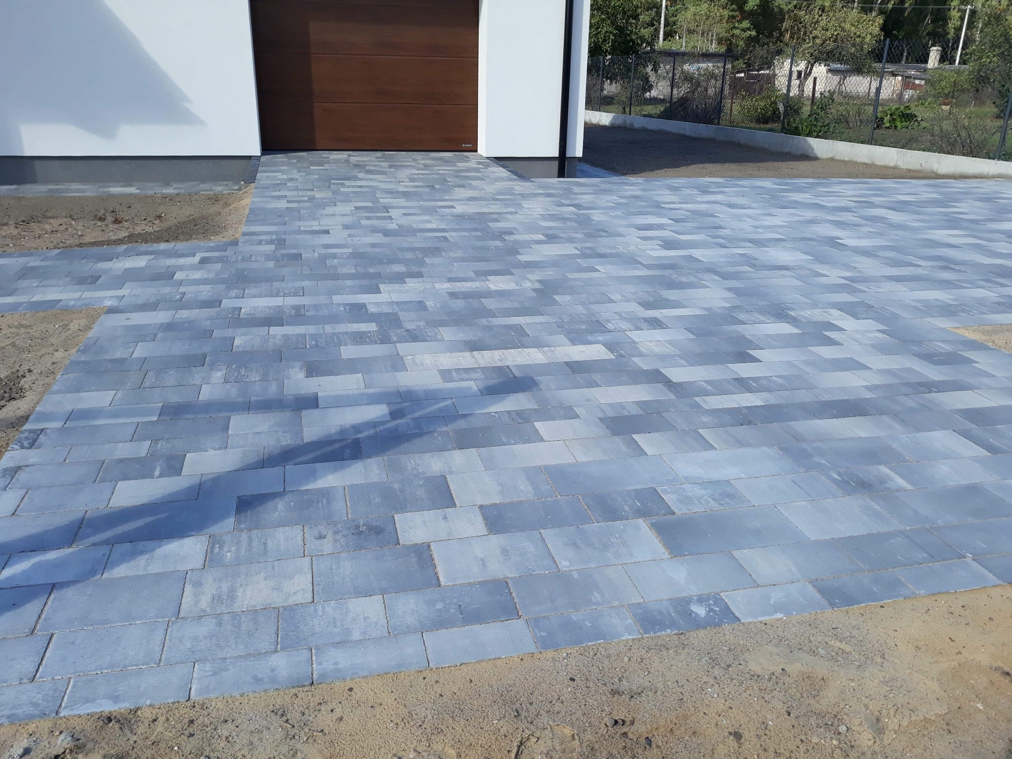 Nowa, szara kostka brukowa ułożona przed nowoczesnym domem z brązową bramą garażową. Widoczne niedociągnięcia na obrzeżach i piasek.