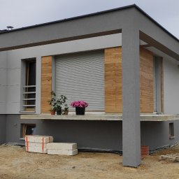 zrbmalmar - Nowoczesny dom jednorodzinny z szarą elewacją, drewnianymi panelami, roletami zewnętrznymi, kwiatami w donicach na balkonie i widocznymi fundamentami w trakcie prac wykończeniowych.