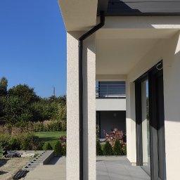 zrbmalmar - Nowoczesny dom z jasną elewacją o fakturze baranka i ciemnoszarą stolarką okienną oraz rynnami. Widoczny fragment tarasu wyłożonego szarymi płytami i przygotowany teren pod dalsze prace ogrodowe.