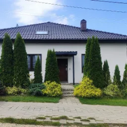 Nowoczesny dom jednorodzinny z białą elewacją, ciemną dachówką i symetrycznie posadzonymi tujami, widok z chodnika, biały bus zaparkowany z boku.