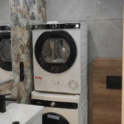 Nowoczesna łazienka z pralką i suszarką w kolumnie, umywalką z szafką oraz toaletą, wykończona płytkami imitującymi kamień i drewno.